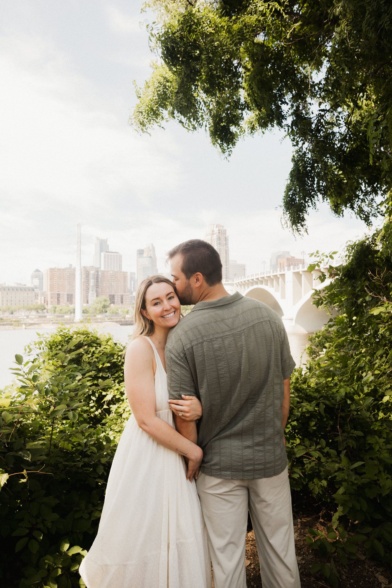 Couple embracing outdoors near a river with city skyline and bridge in background, woman in white dress smiling.