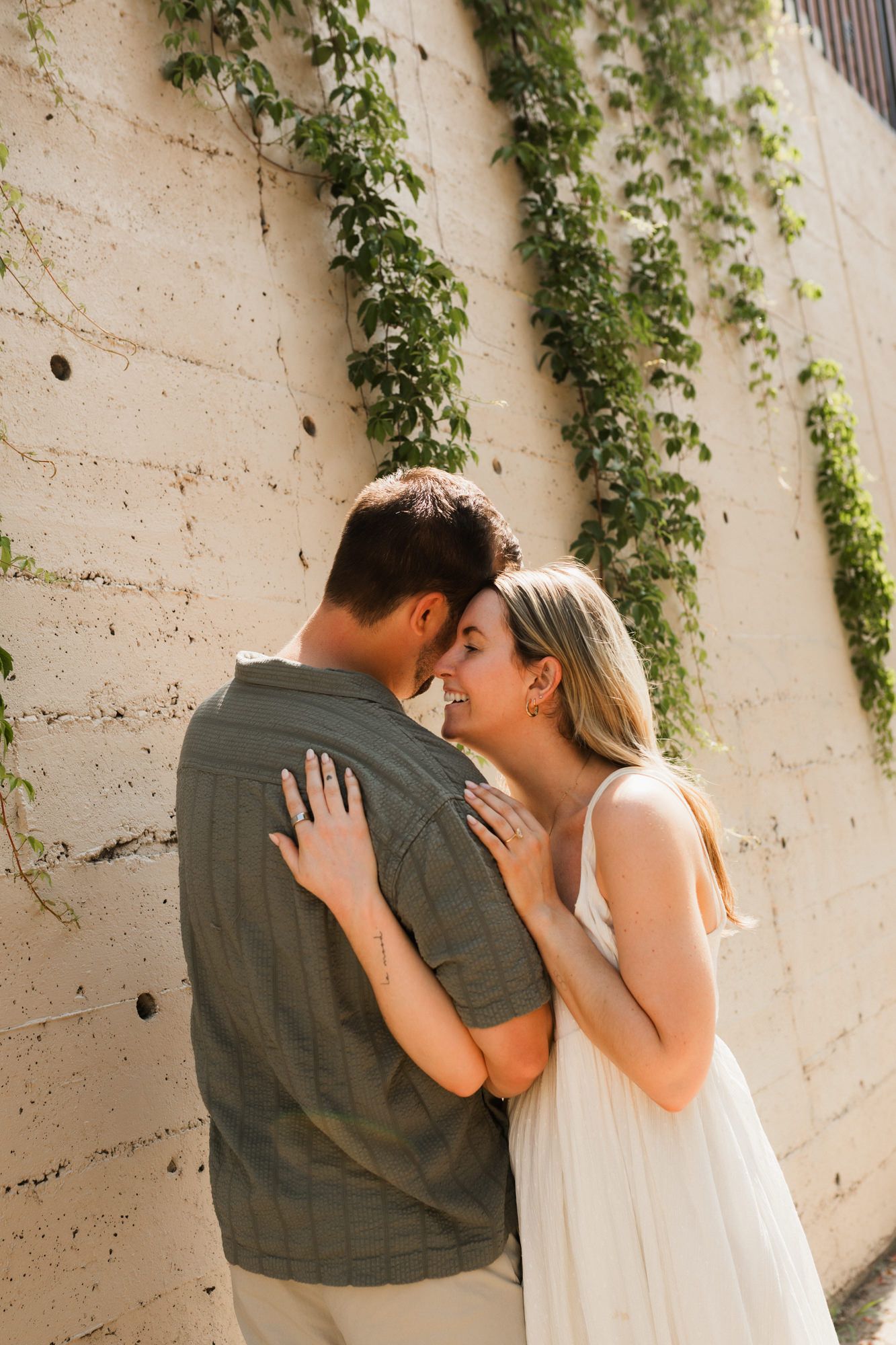 Couple sharing a romantic moment against a vine-covered wall, woman in white dress touching man's back.