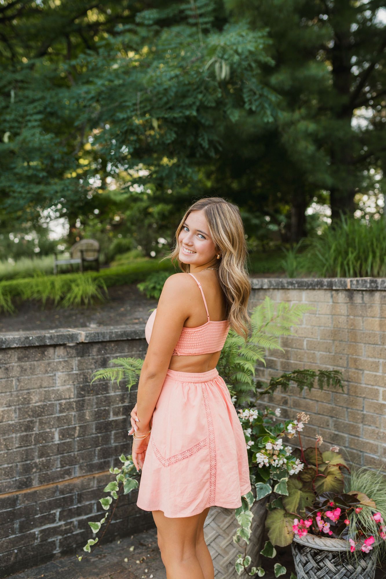 Young woman in pink crop top and skirt smiling over shoulder in lush garden setting.