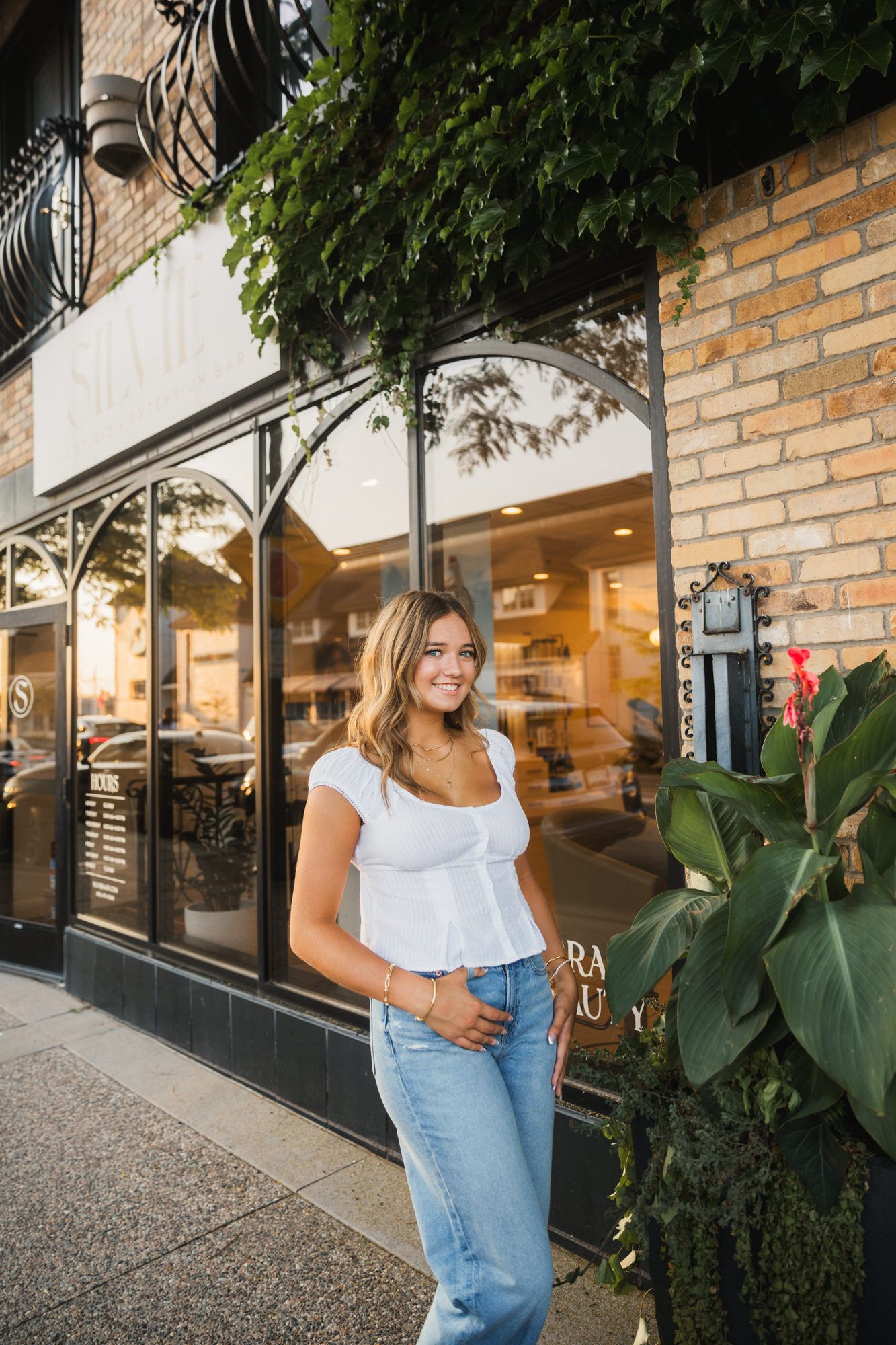 Smiling young woman in white top and jeans posing outside a brick storefront with lush greenery.