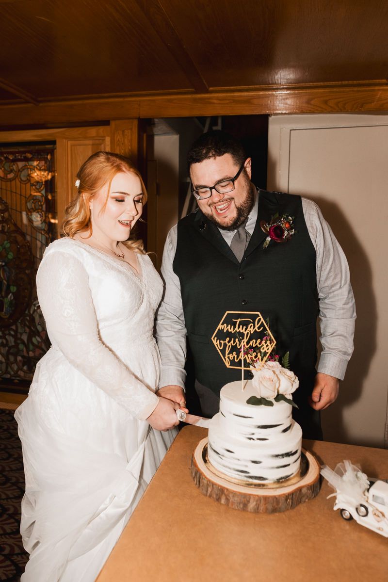 Couple cutting their elegant white wedding cake together on a wooden display stand during their reception celebration.