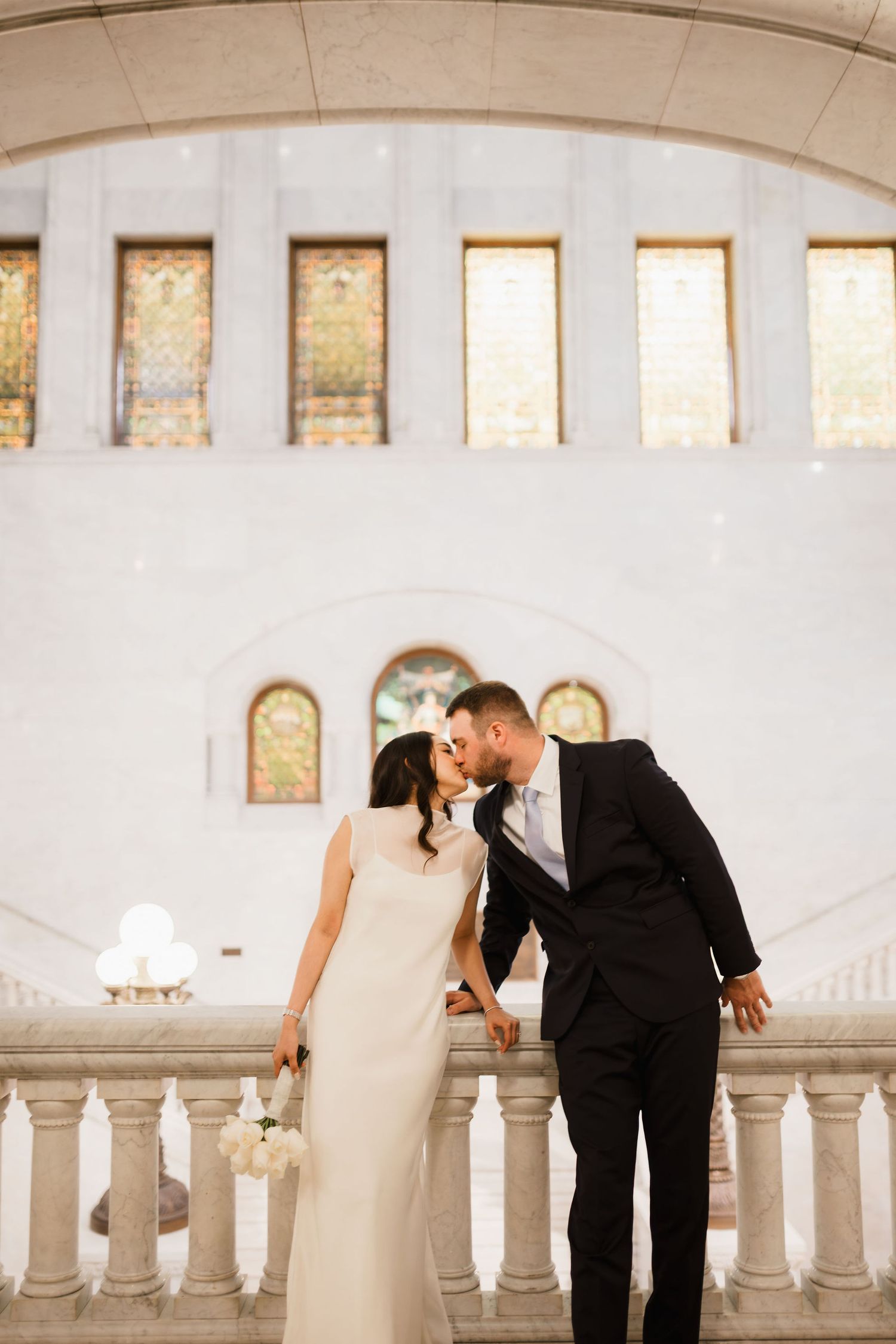 Bride and groom share a romantic kiss on an elegant balcony inside a grand white wedding venue.