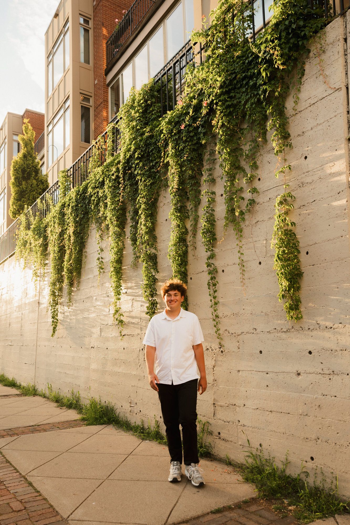 Young person in white shirt and black pants walking along a sunlit sidewalk beside an ivy-covered white brick wall.