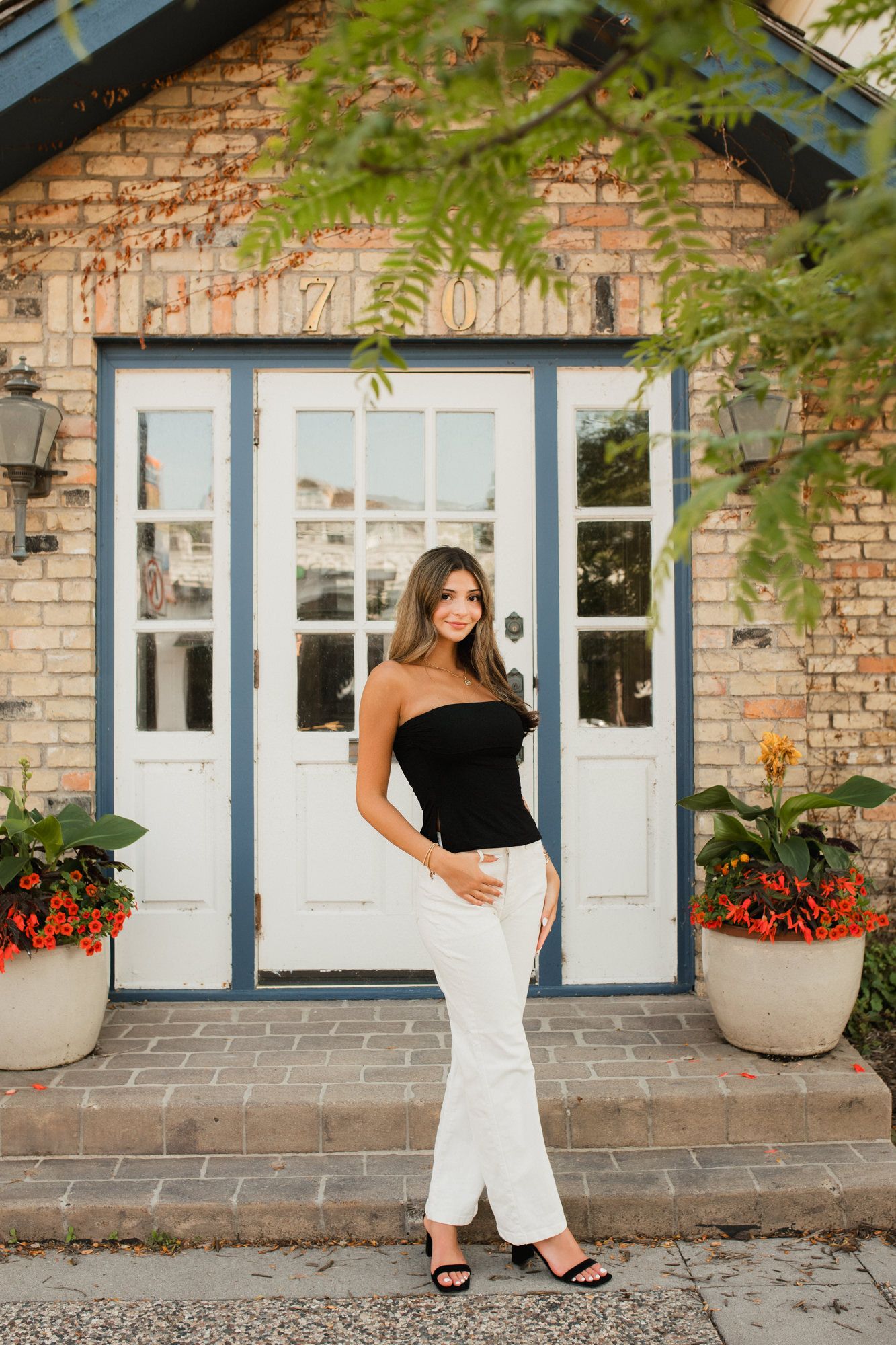 Young woman in black tube top and white pants posing in front of a brick building with white French doors.