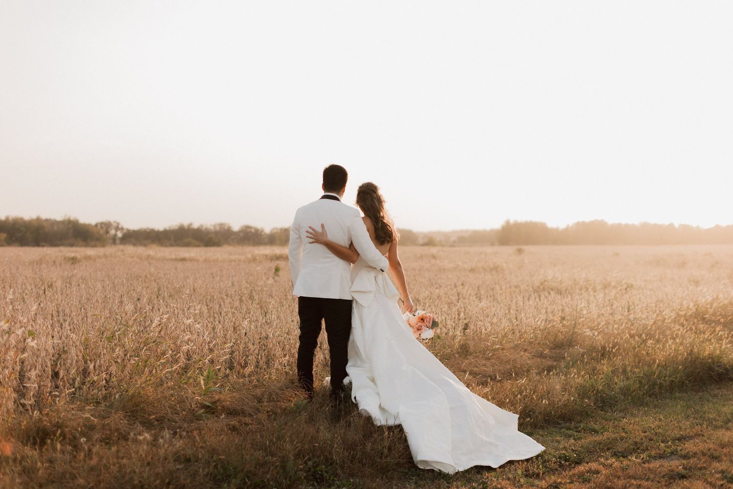 A bride and groom stand together in a golden field at sunset, the bride holding a bouquet in her flowing white gown.