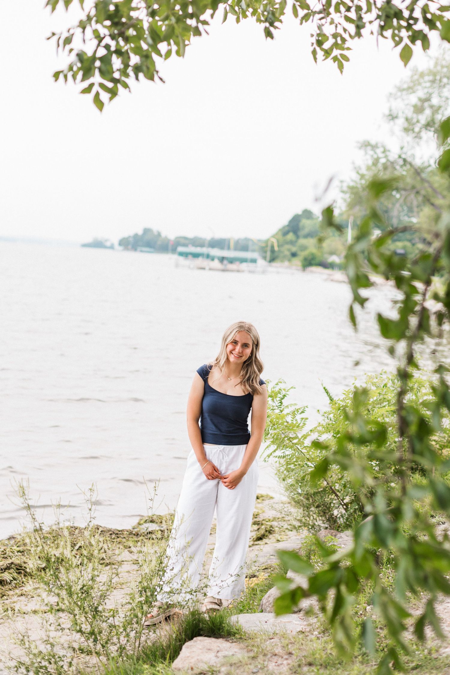 A person in white pants and navy top walks along a rocky beach shoreline framed by green leaves.