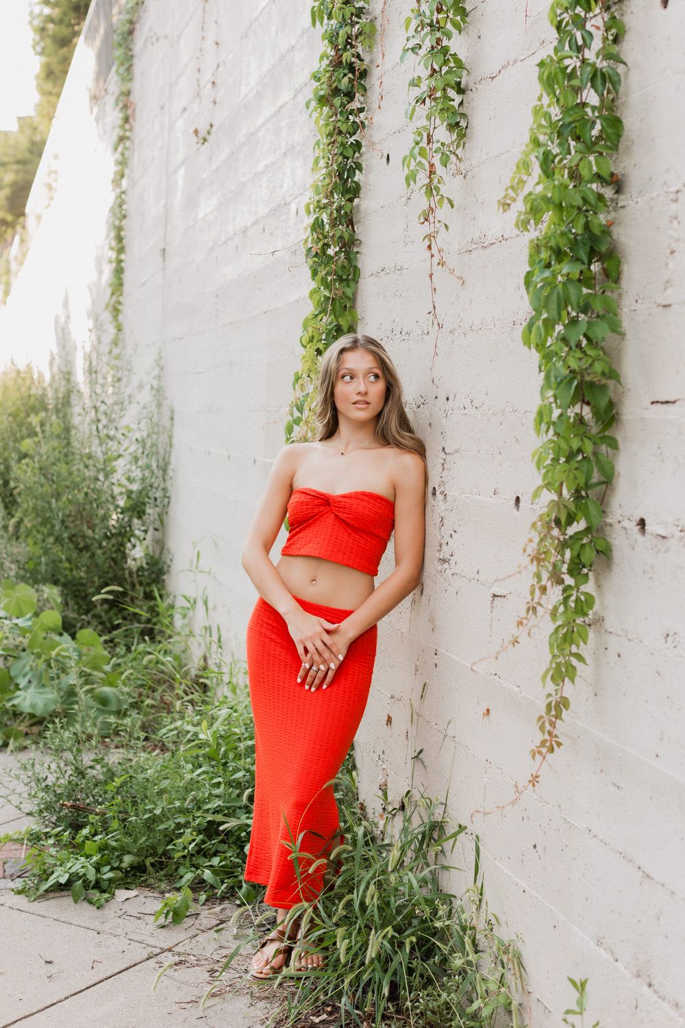 Woman in red dress poses against white wall with hanging vines in outdoor portrait series.