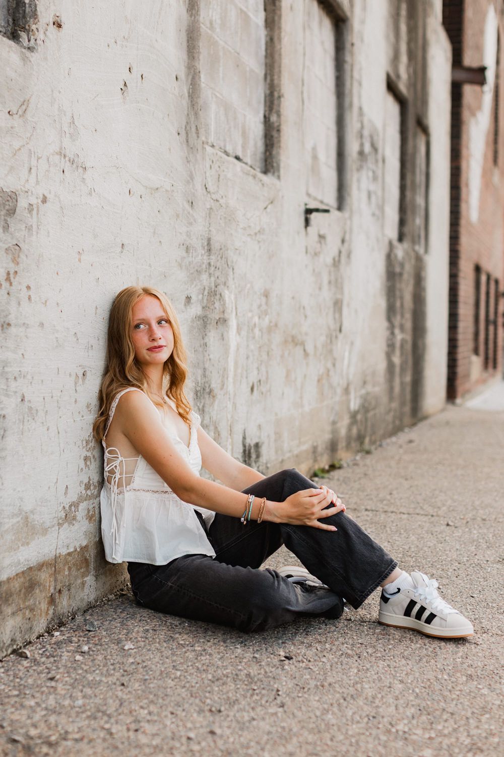 Person in white tank top and black pants sits casually against weathered wall on urban sidewalk wearing Adidas sneakers.