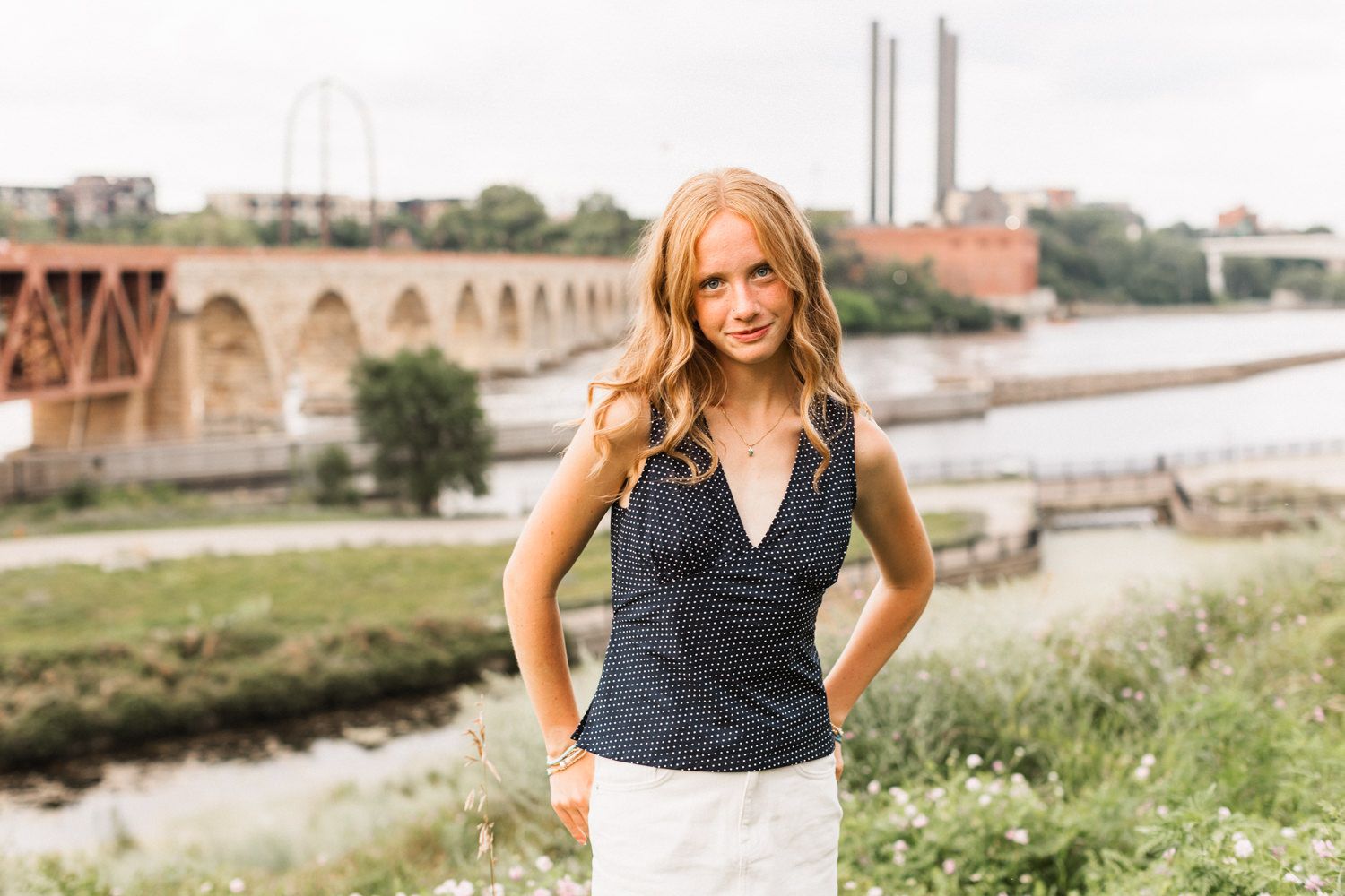 A person in a sleeveless polka dot blouse stands along a riverbank with historic architecture in the background.