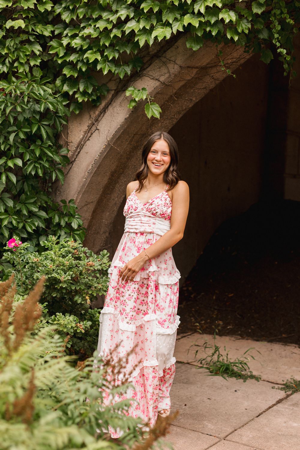 A floral maxi dress is showcased against a stone archway covered in lush green ivy and garden foliage.