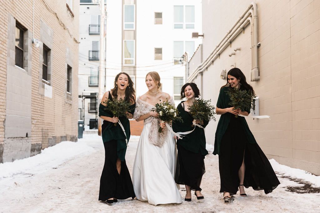 Wedding group stands in formation along a light colored exterior walkway wearing formal black and white attire.