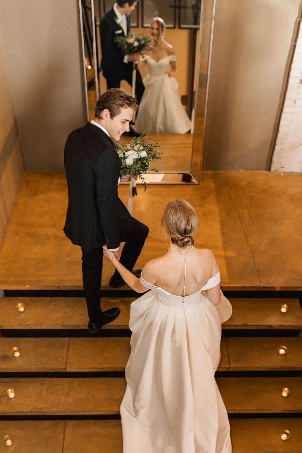 A bride in an elegant off-shoulder gown and groom share tender moments in a warmly lit indoor space.