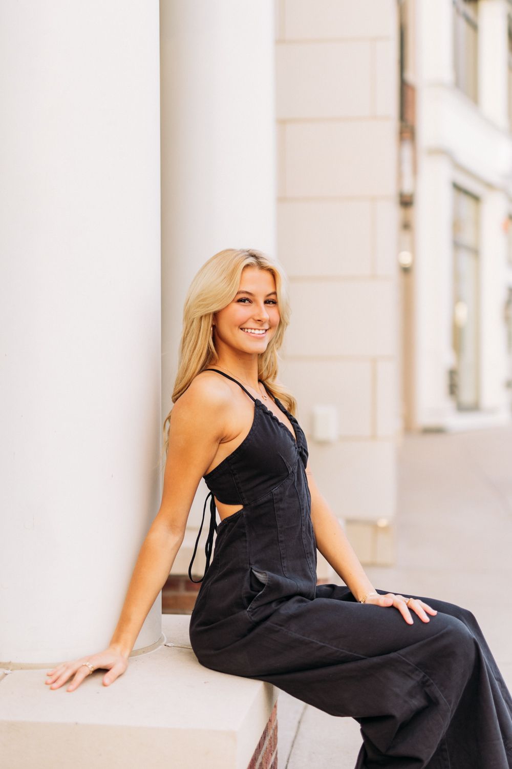 A woman in a elegant black jumpsuit poses gracefully on outdoor steps near white columns.