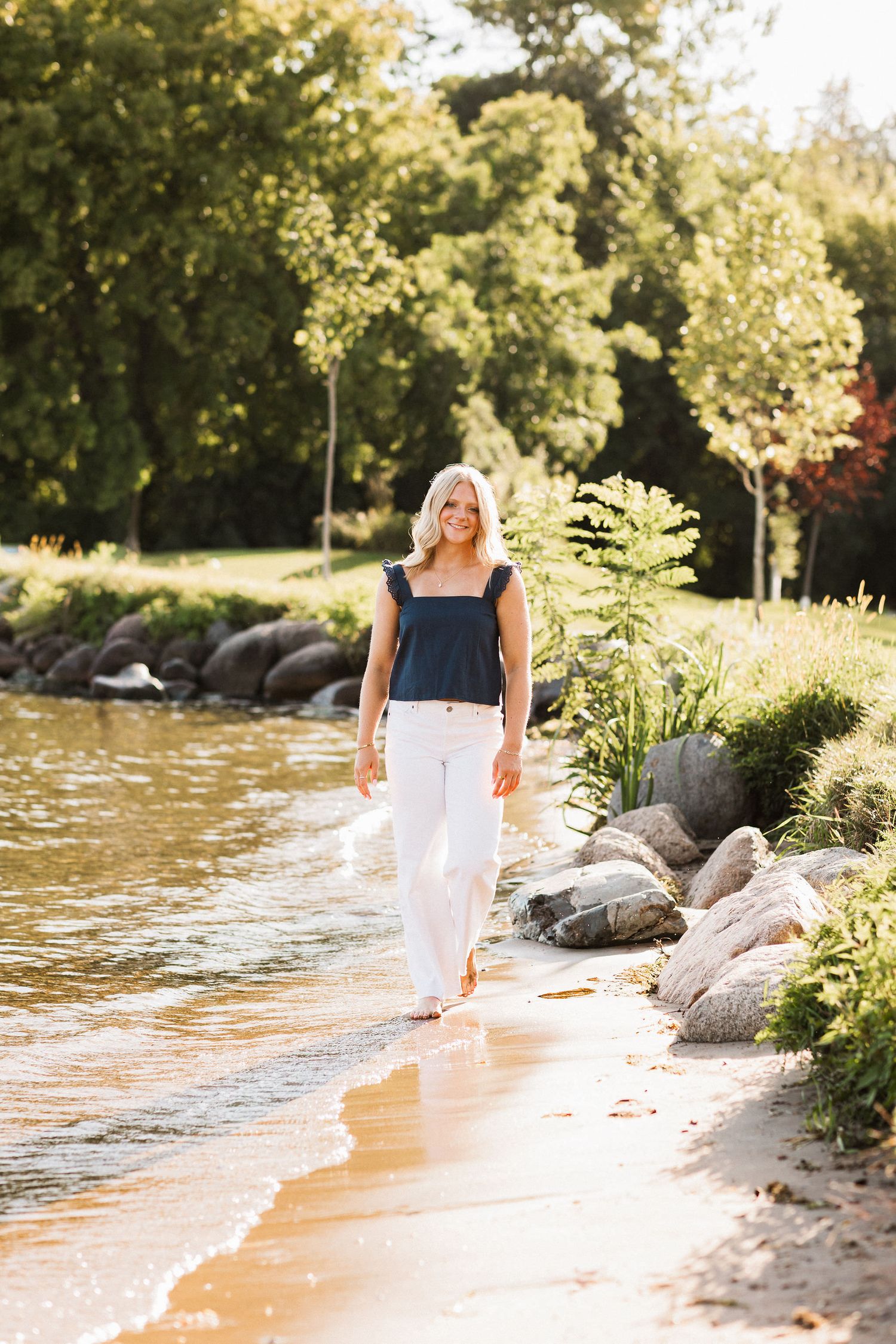 Figure in a white dress walks along a serene lakeside beach during golden hour with rocks and foliage in view.