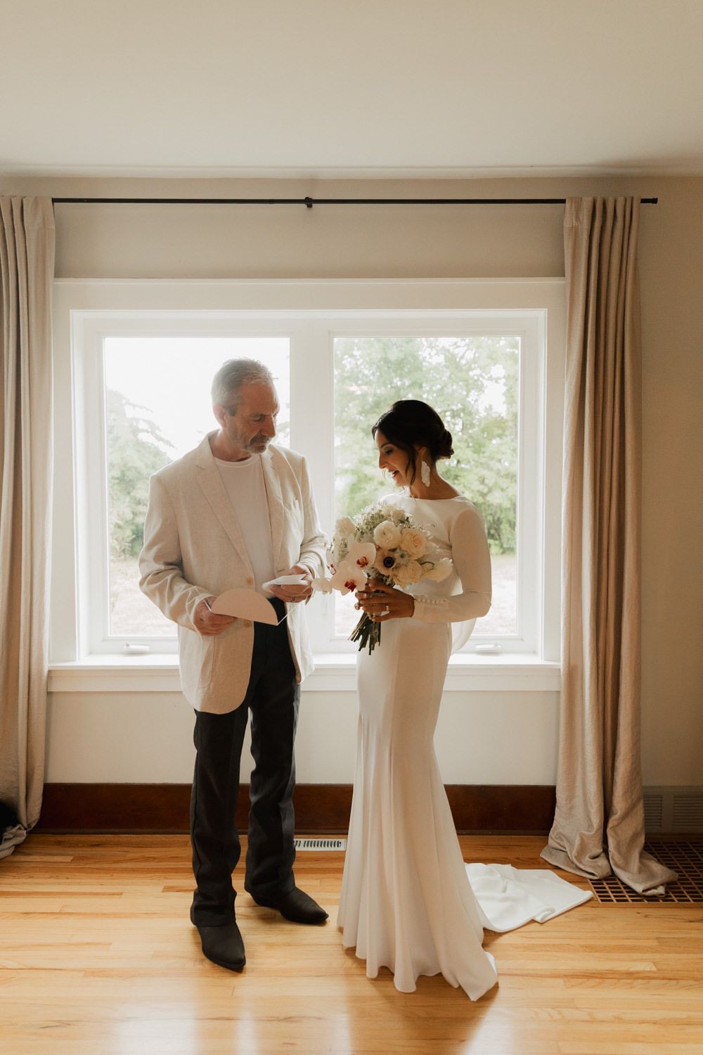 A tender moment between two people in formal attire by a bright window, with the bride holding a white floral bouquet.