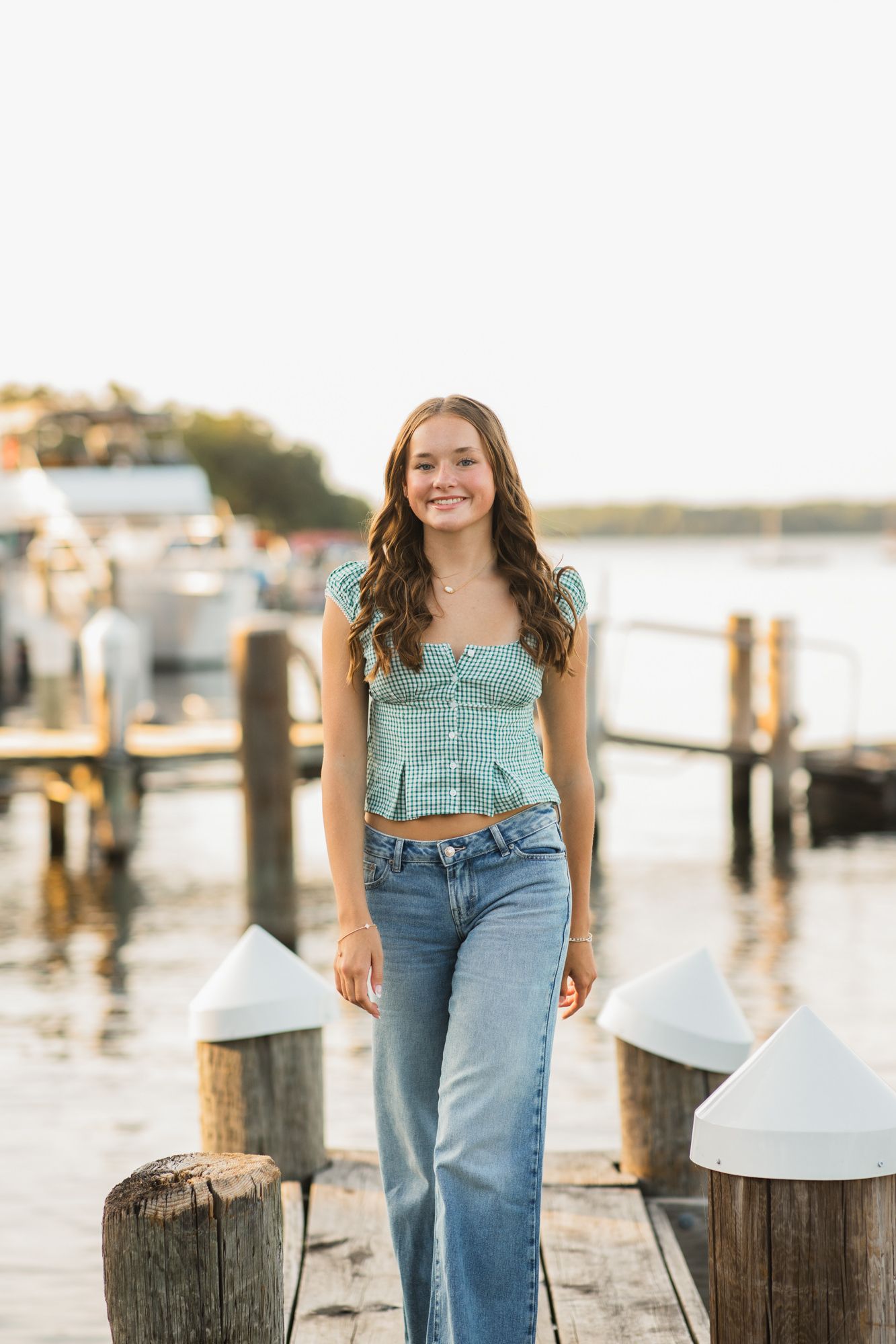 Young woman in green crop top and jeans smiling on a wooden dock by the water at golden hour.