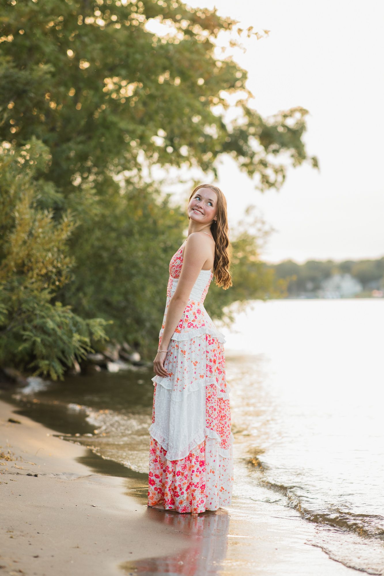 Young woman in floral maxi dress standing barefoot on sandy beach shore at golden hour sunset.