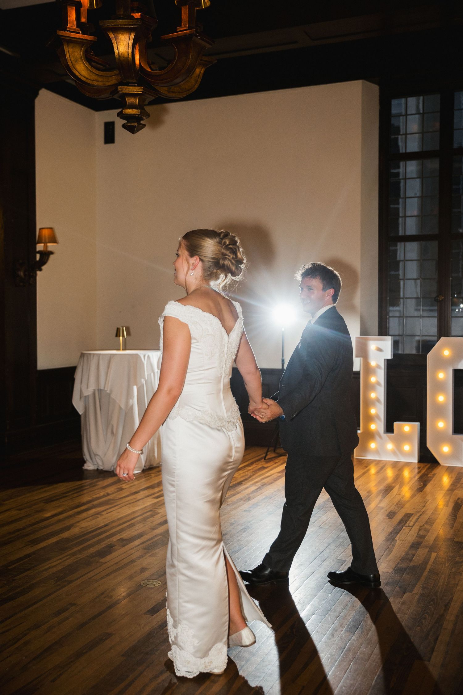 Bride and groom walk hand in hand toward illuminated LOVE letters on a warmly lit wedding reception dance floor.