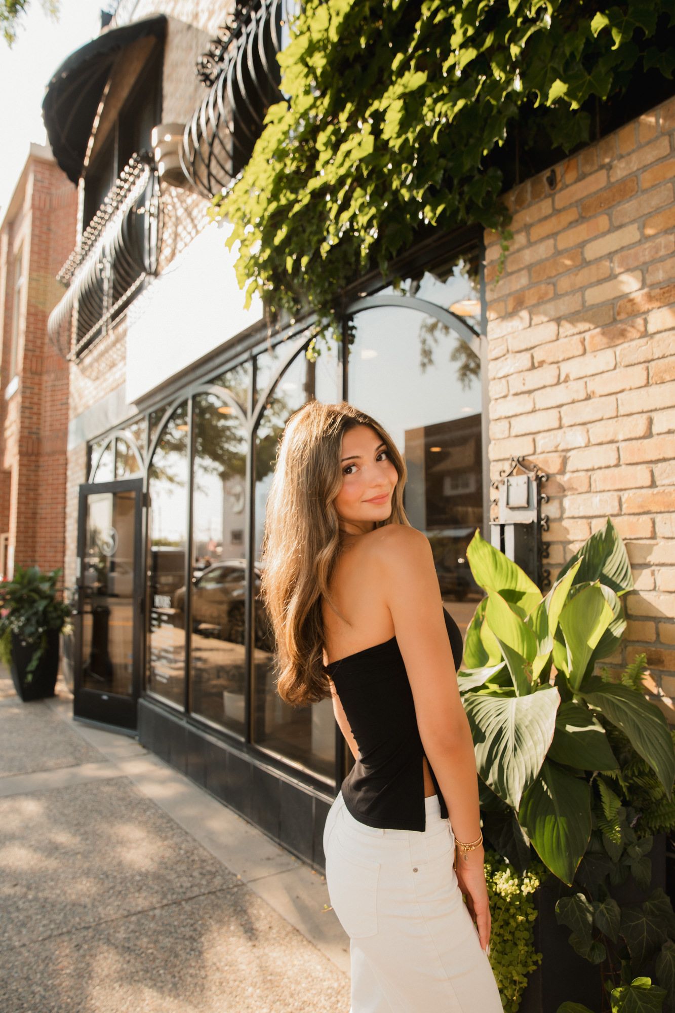 Young woman in black top and white skirt posing outside a brick building with lush green plants.