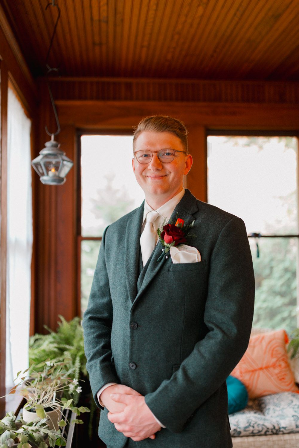 A groom in a dark suit and glasses smiles warmly in a wood-paneled room during a wedding celebration.