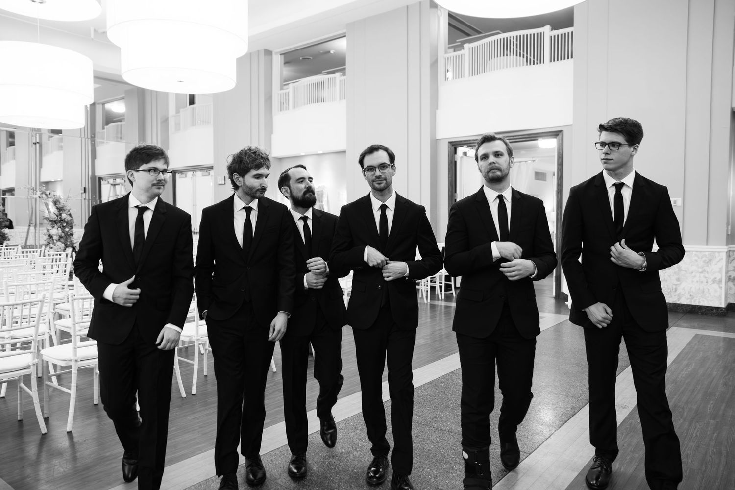 Six men in black suits walking together in a bright venue in a black and white wedding photo.