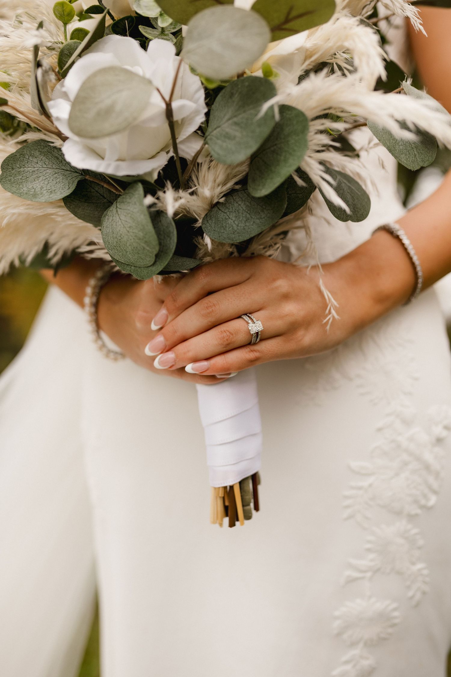Bride holding boho wedding bouquet with white roses, eucalyptus, and pampas grass, wearing diamond ring.