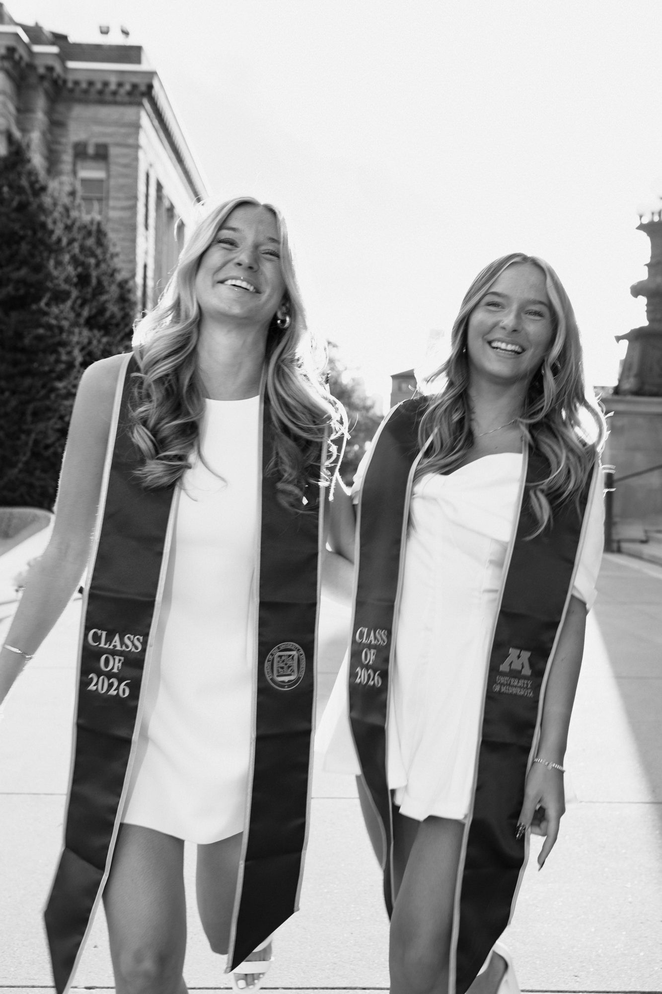 Two smiling University of Minnesota Class of 2026 graduates in white dresses and black stoles walking on campus.