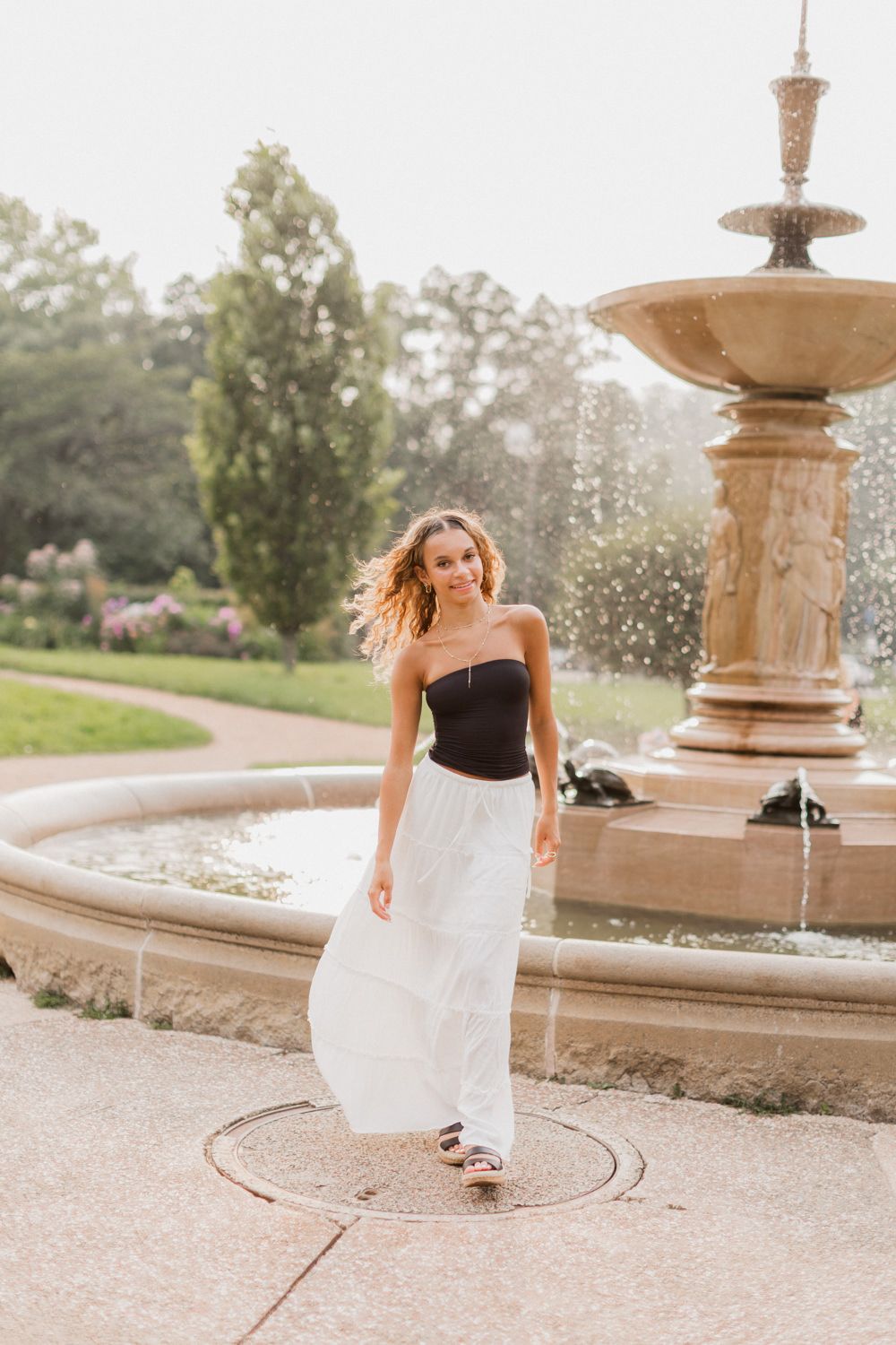 A woman in a black top and white skirt spins gracefully near an ornate stone fountain in a garden setting.