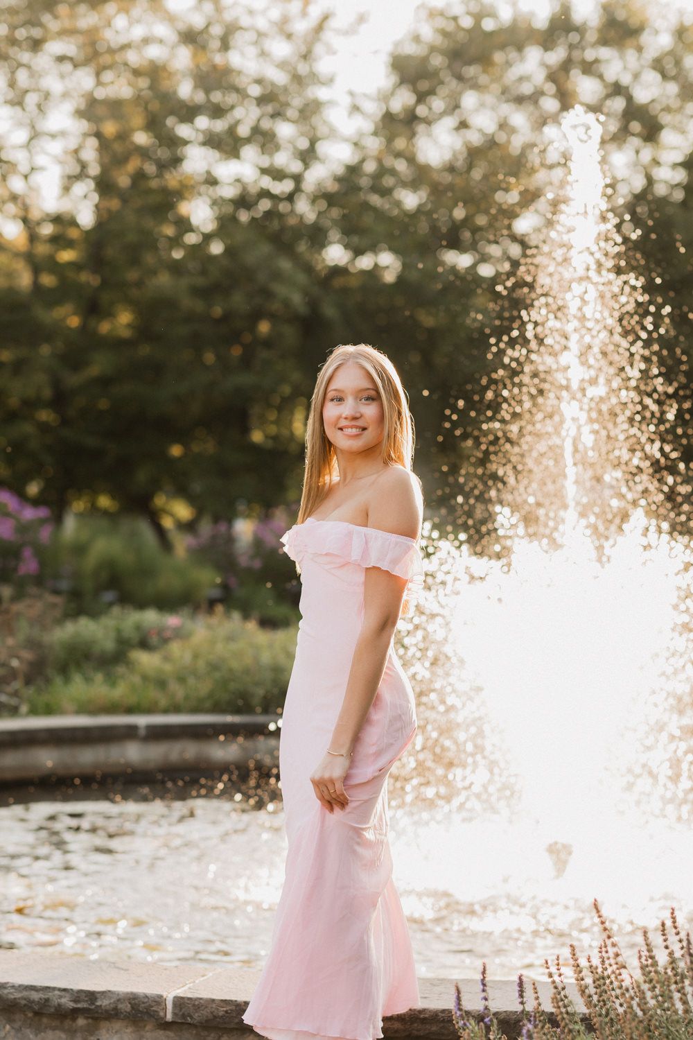 A person in a pink dress stands near a fountain at sunset in a garden setting.