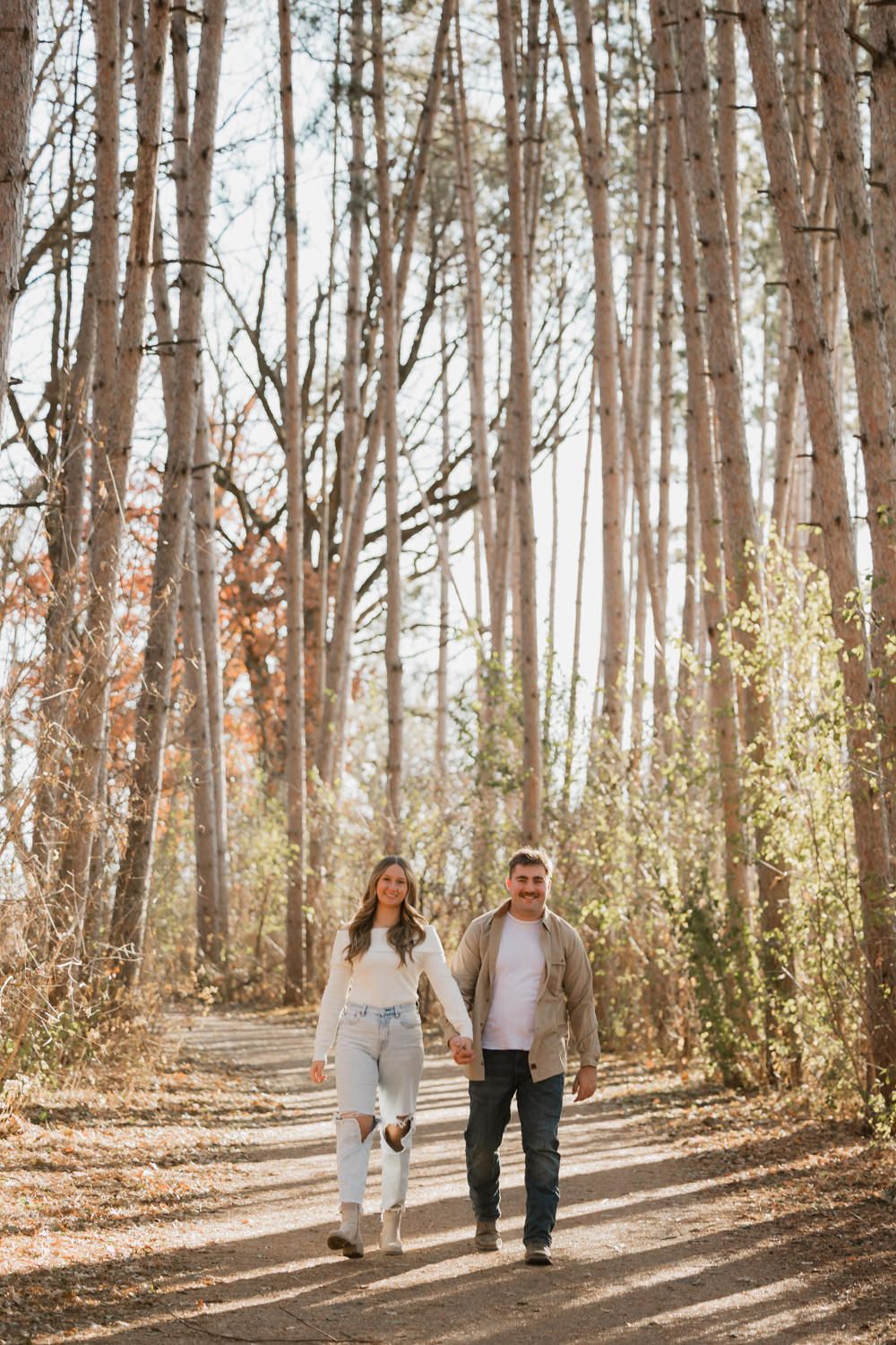 Couple walking hand in hand through a sunlit forest path with tall trees casting long shadows on the ground.
