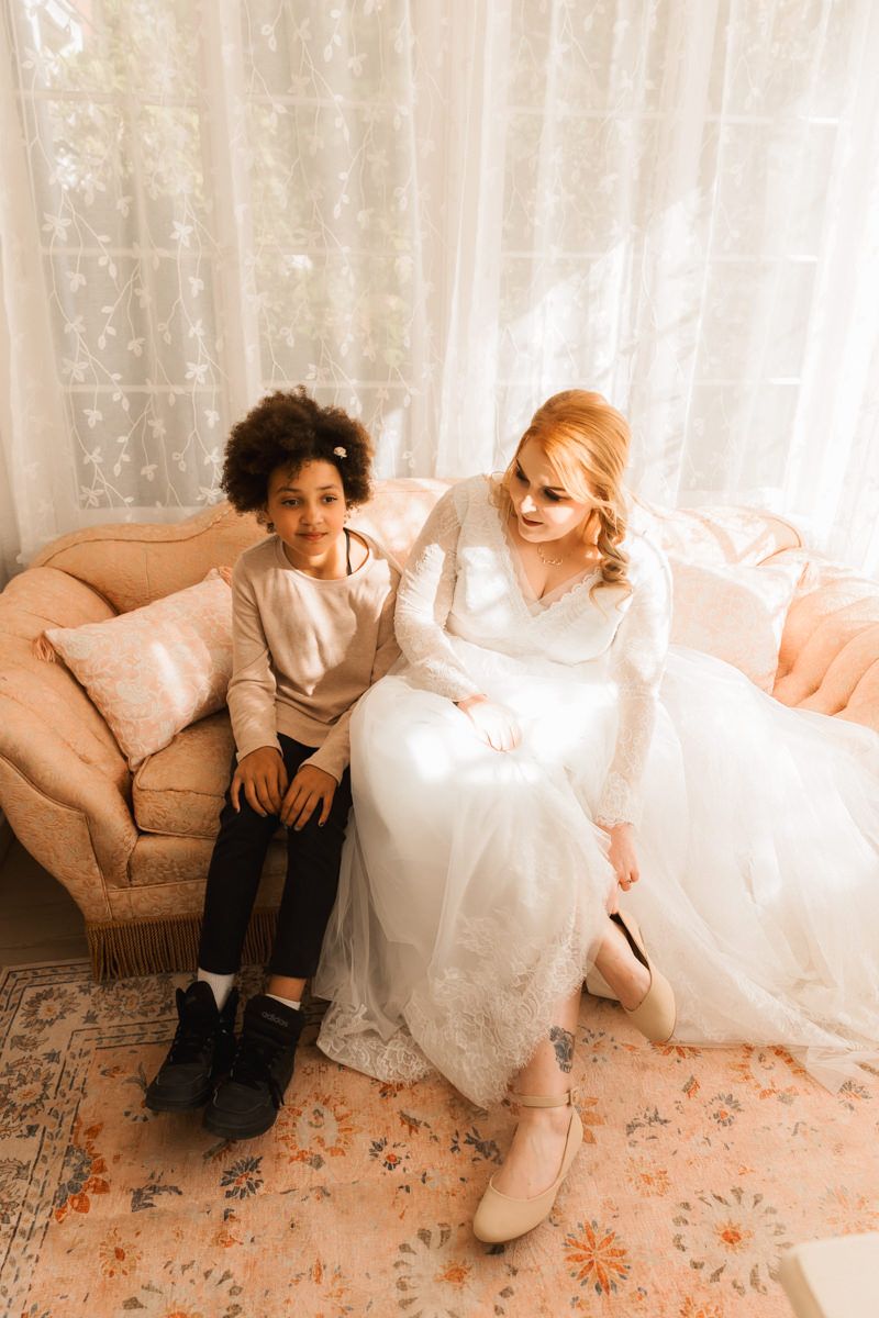 Two people in wedding attire sit casually on the floor against white curtains and floral carpet.