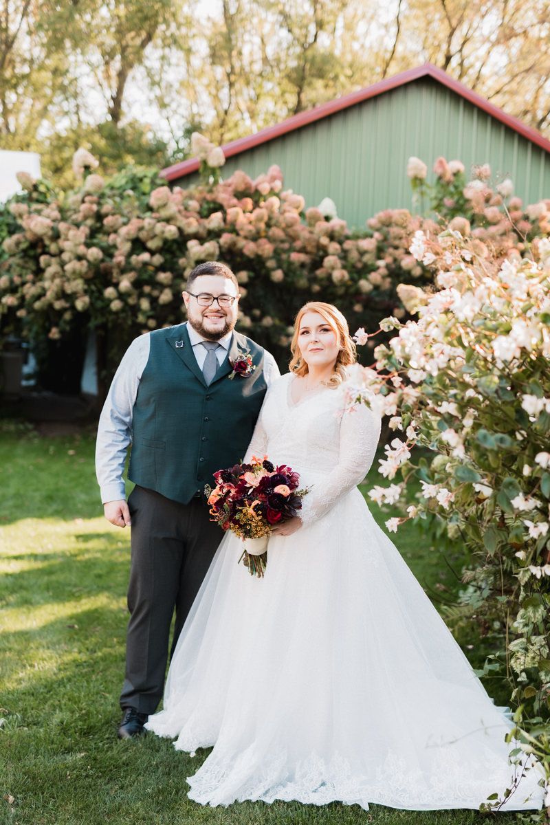 A couple poses together in their wedding attire against a backdrop of blooming rose bushes and a green barn structure.