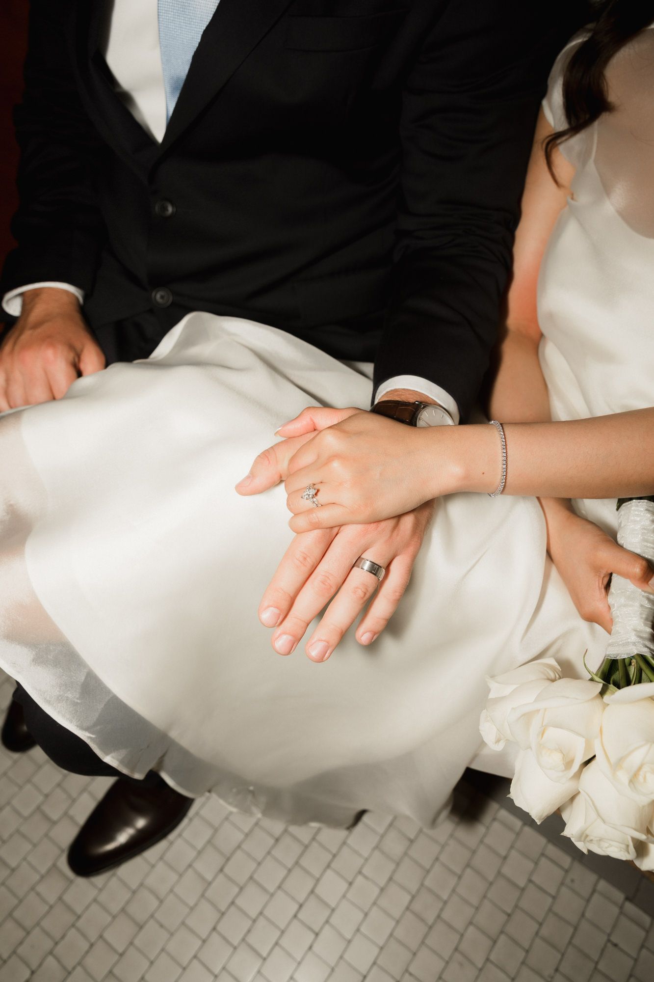 Newlyweds holding hands showing wedding rings, bride in white dress with bouquet of white roses.