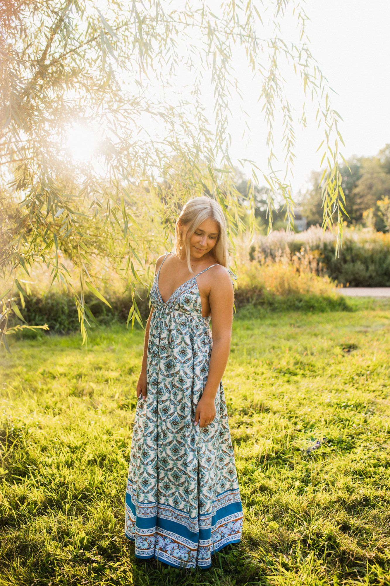 Woman in blue floral maxi dress standing in sunlit garden with willow tree and green grass backdrop.