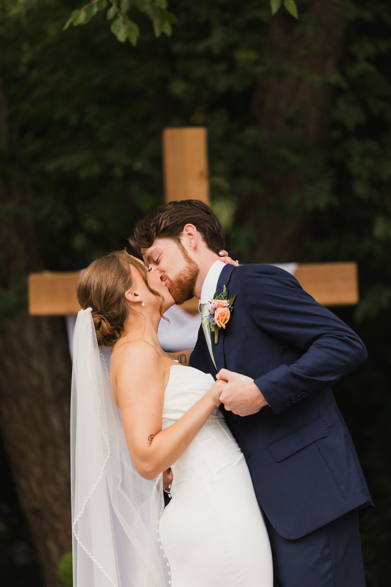 Bride and groom share first kiss in front of wooden cross outdoors, bride in white gown with veil, groom in navy suit.