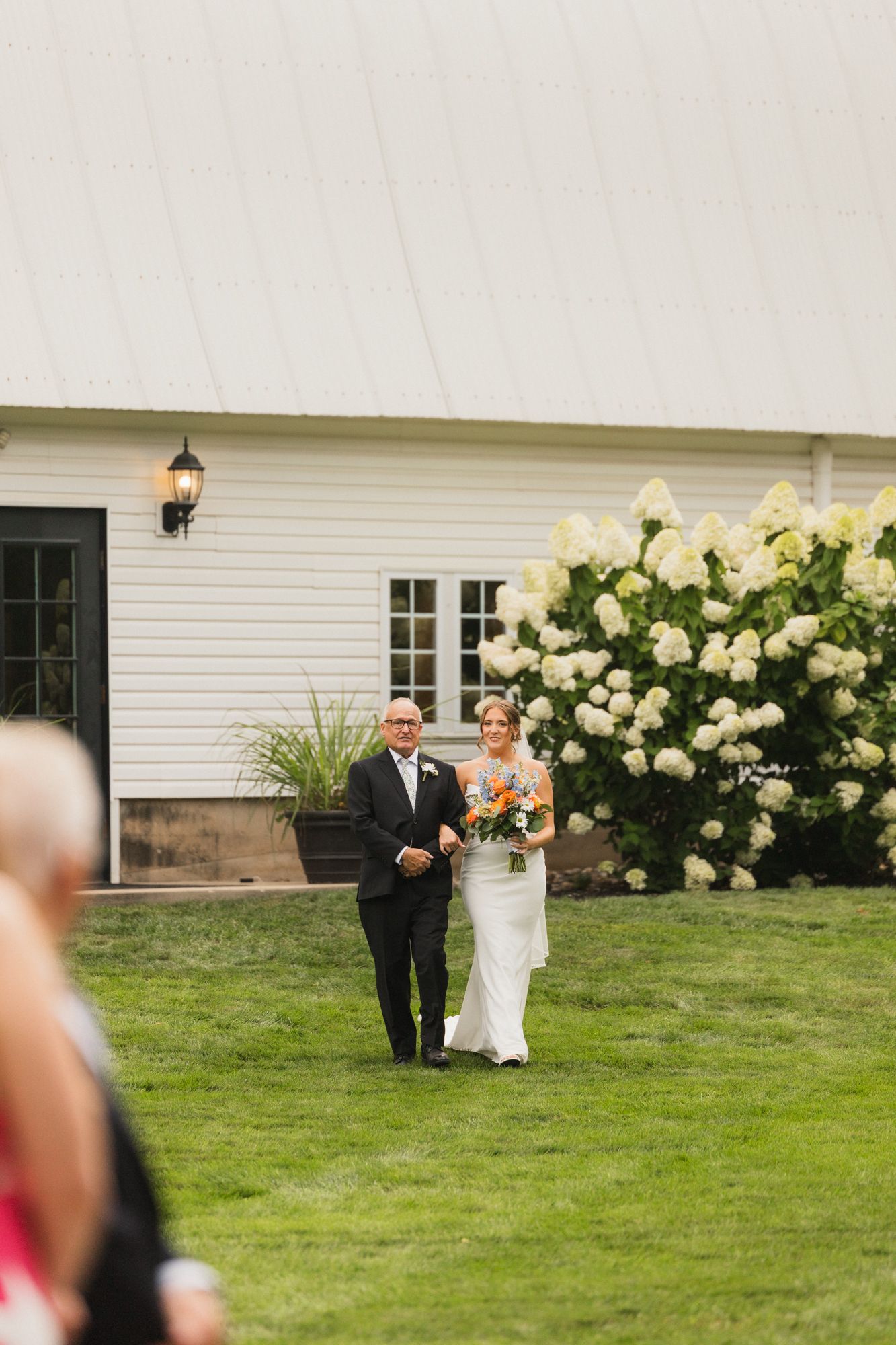 Bride walks down outdoor aisle with escort past white hydrangeas at elegant barn wedding venue.