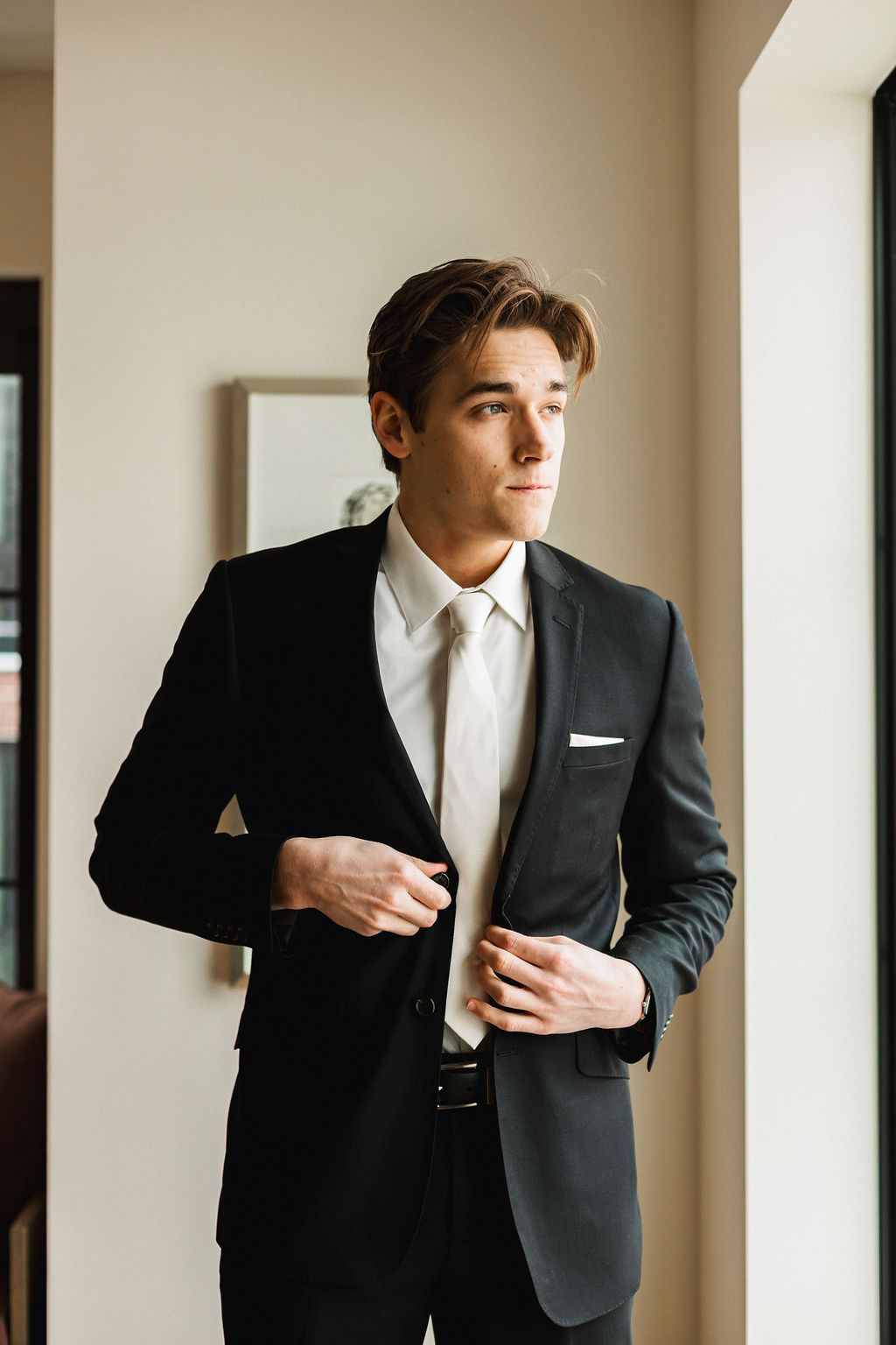 A groom in a classic black tuxedo and white tie adjusts his sleeve in a sunlit room.