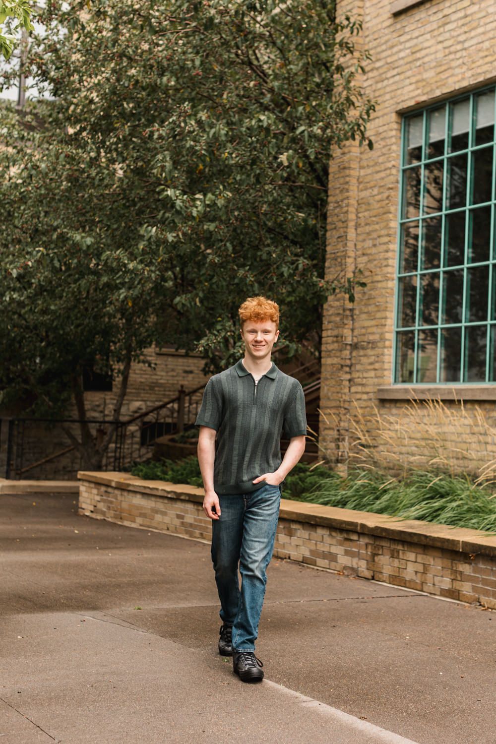 Young person in casual attire standing by a brick building with large windows on a sunny day.