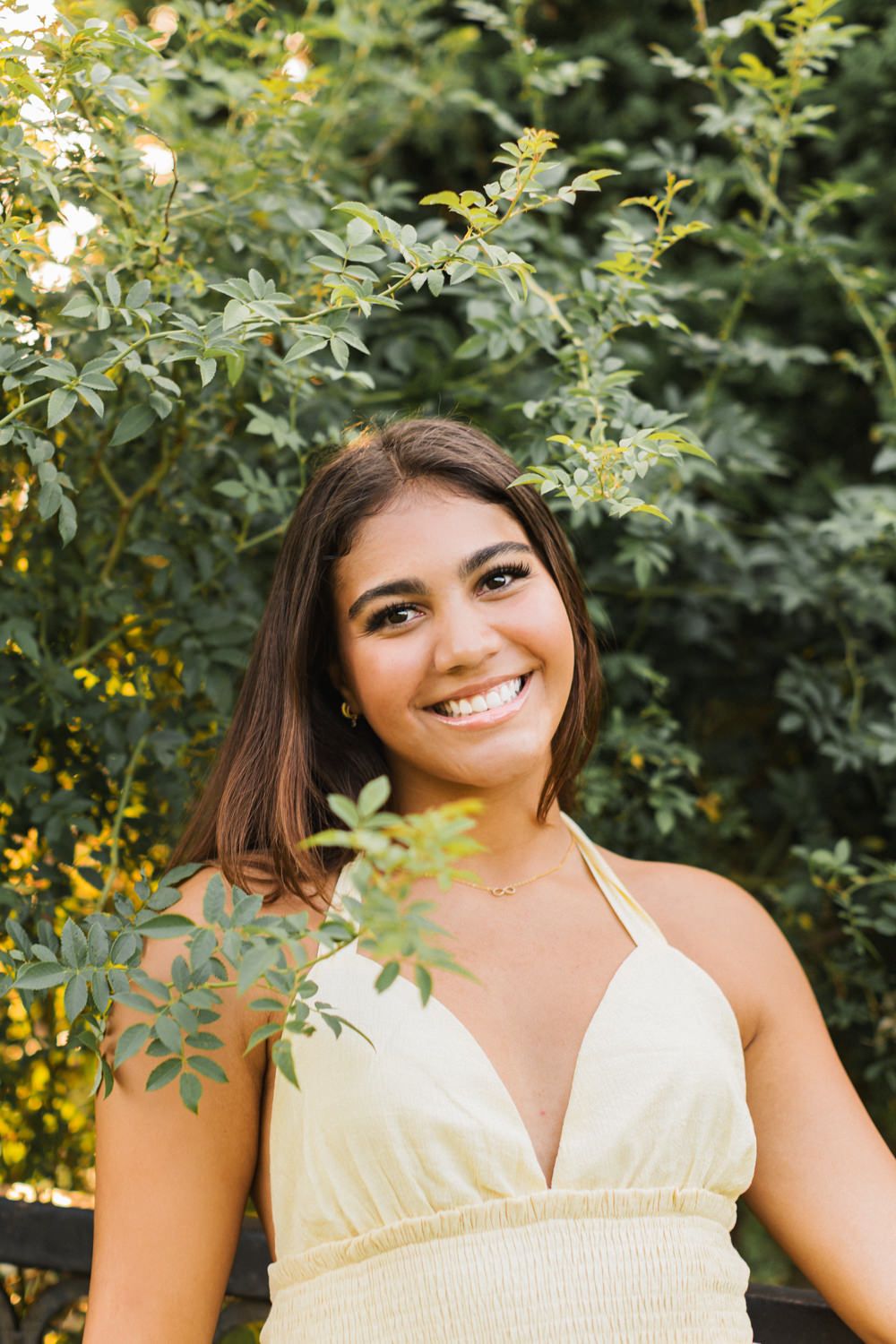 Portrait of a smiling figure in a white dress standing against lush summer foliage in natural outdoor lighting.