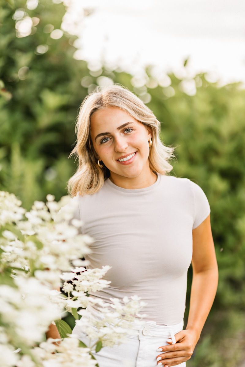 A person in a white t-shirt and light jeans poses outdoors in a sunny garden setting with white flowers.