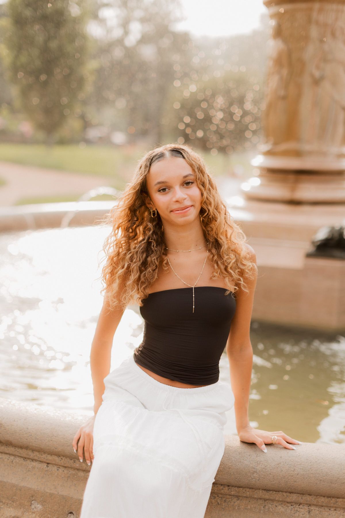 Young woman with curly hair in black tube top and white skirt sitting by a park fountain.