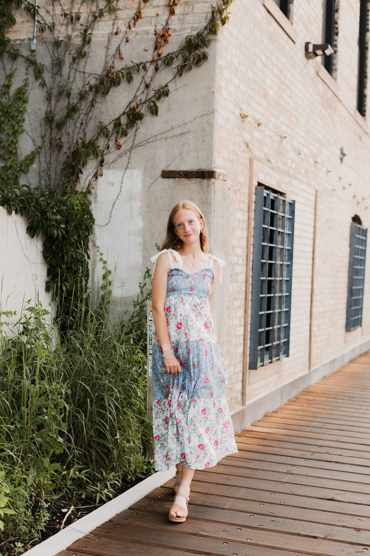 Young blonde woman in blue floral midi dress stands on wooden walkway by brick building.