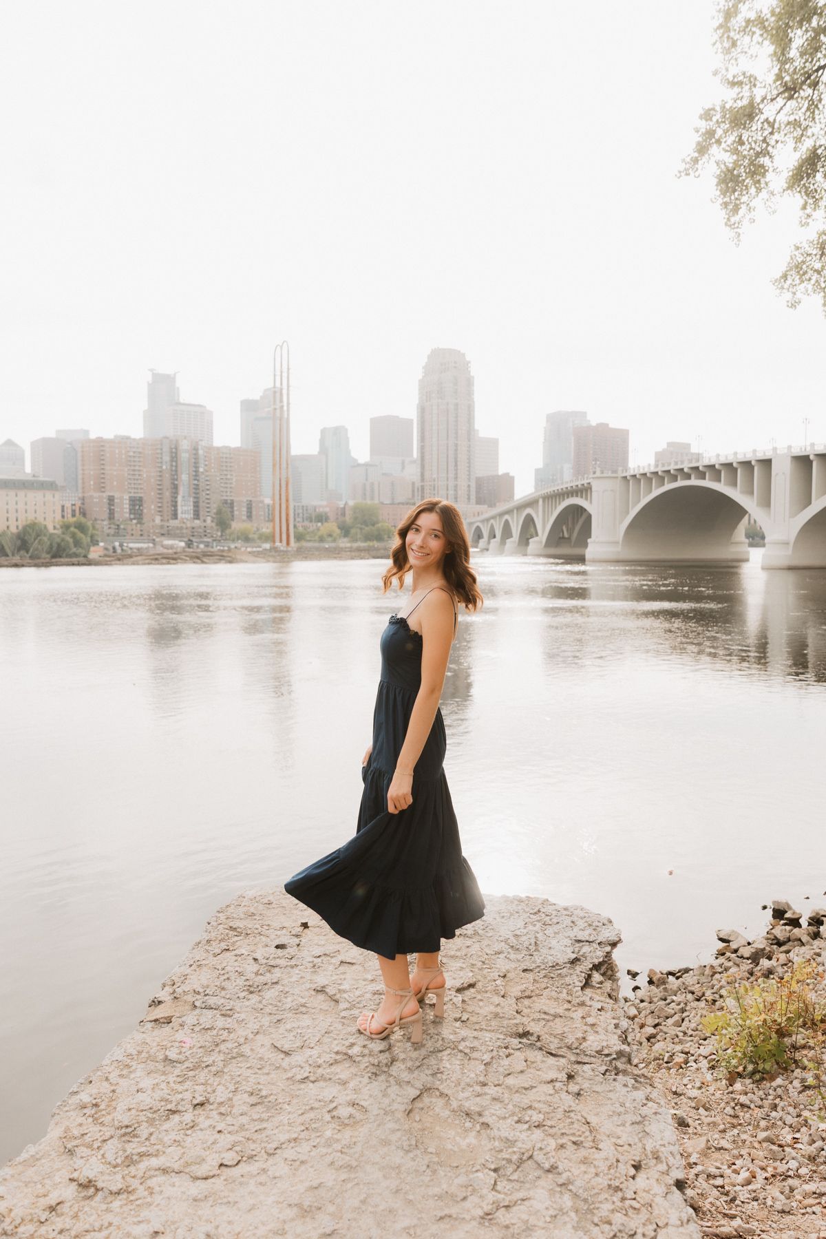 Woman in black midi dress standing on rocky riverbank with city skyline and bridge in background.