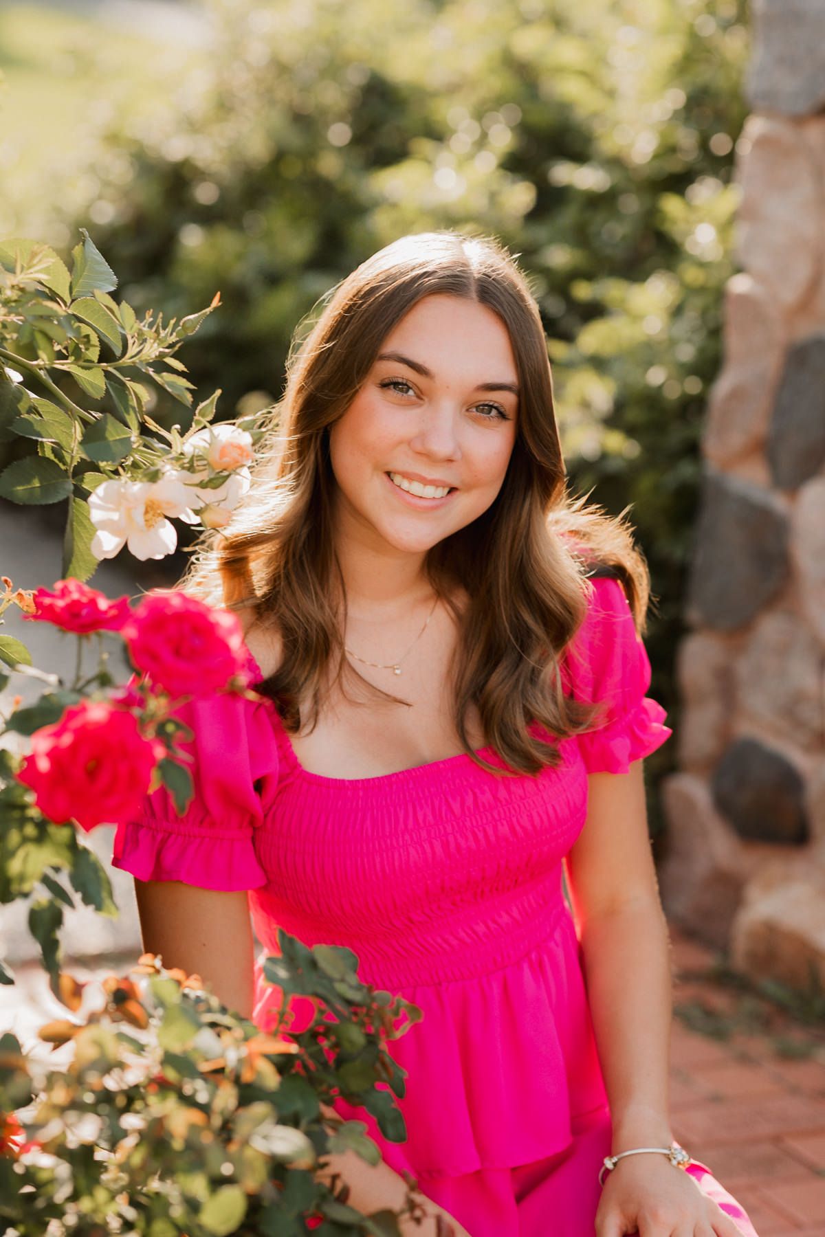Young woman in pink dress smiling among red and white roses in a sunny garden setting.