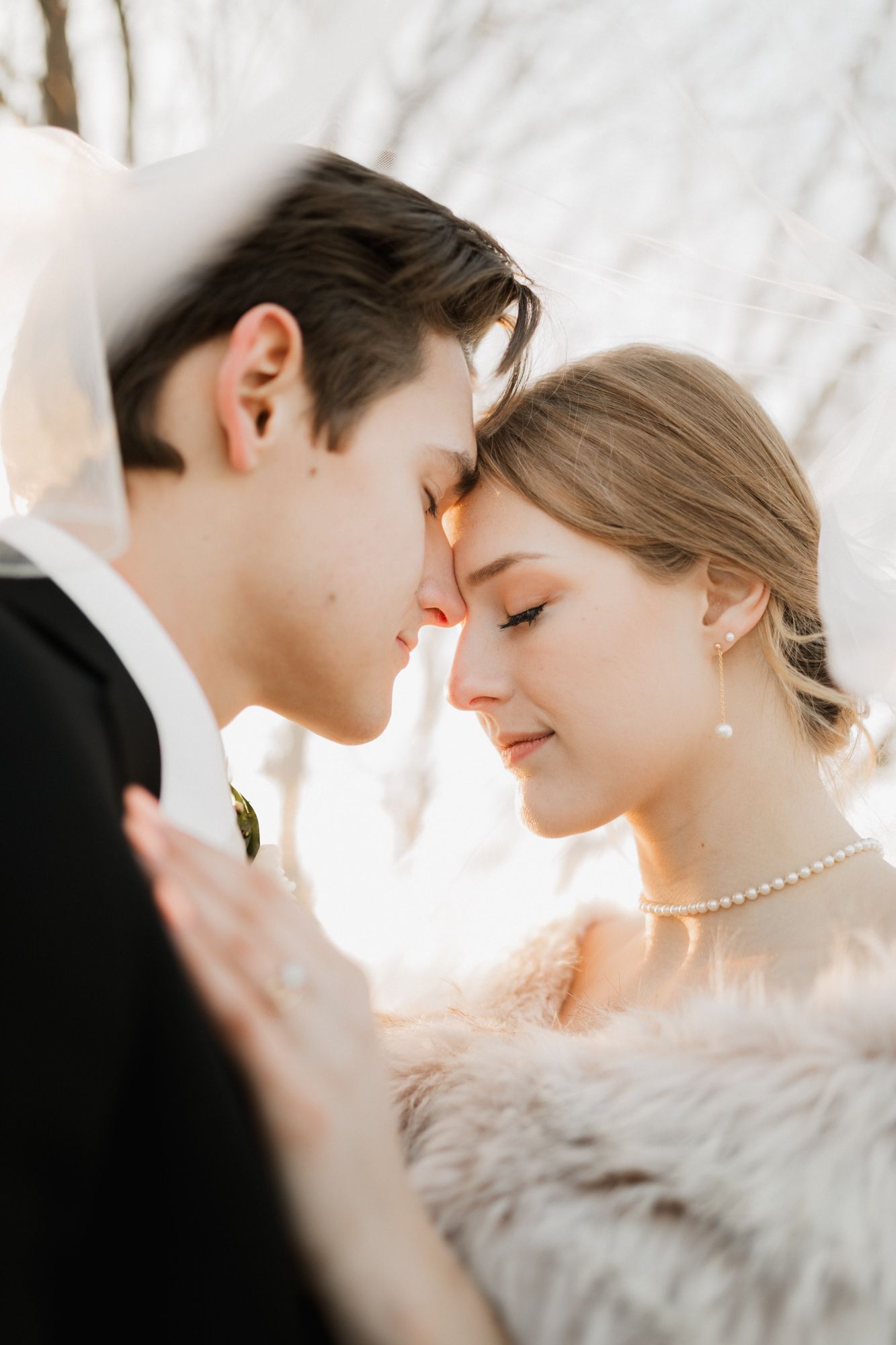 A bride and groom share an intimate forehead touch moment outdoors, bride wearing pearl necklace and fur wrap.
