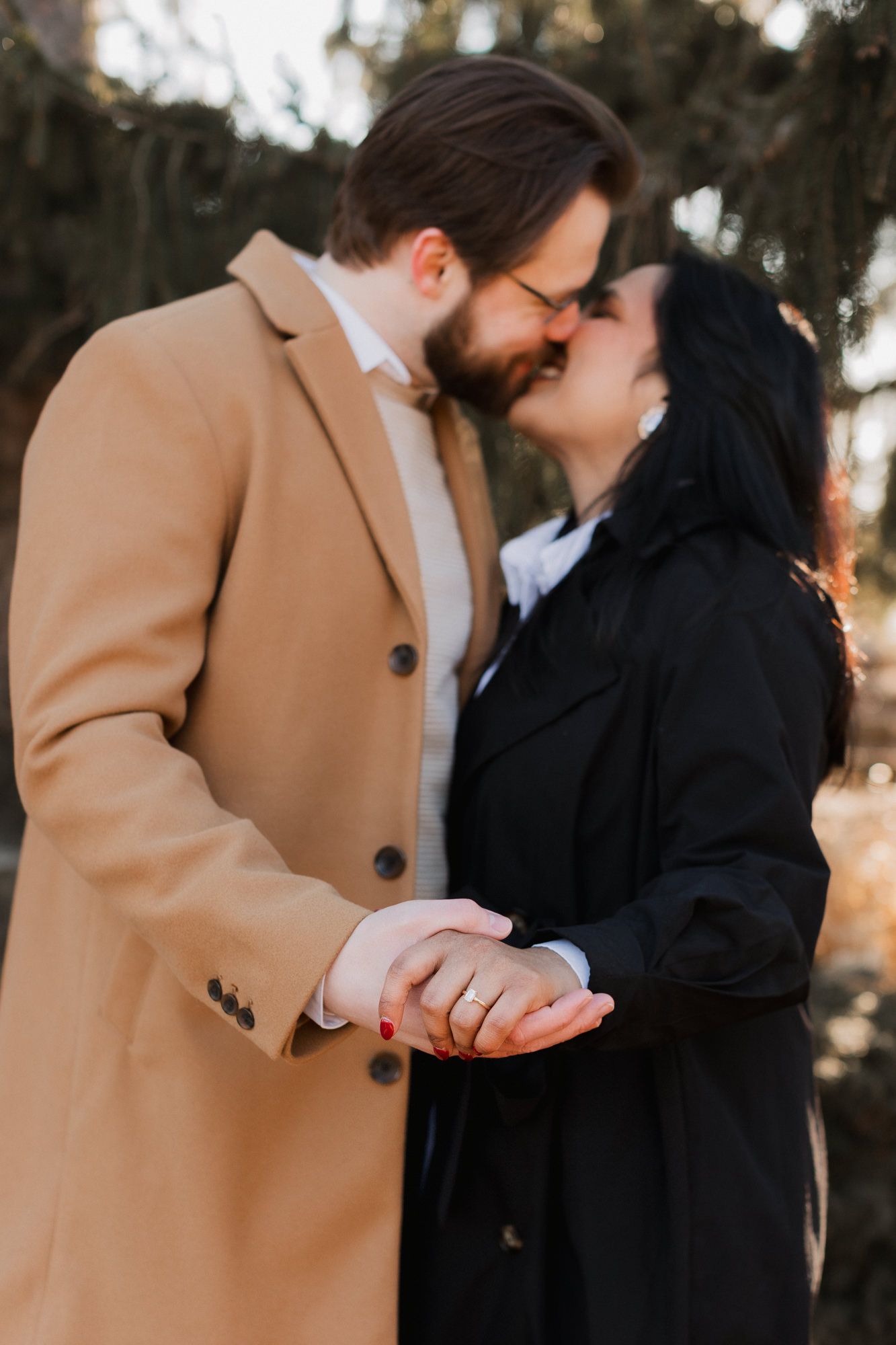 Engaged couple sharing a kiss while holding hands, showcasing a diamond engagement ring in a winter outdoor setting.