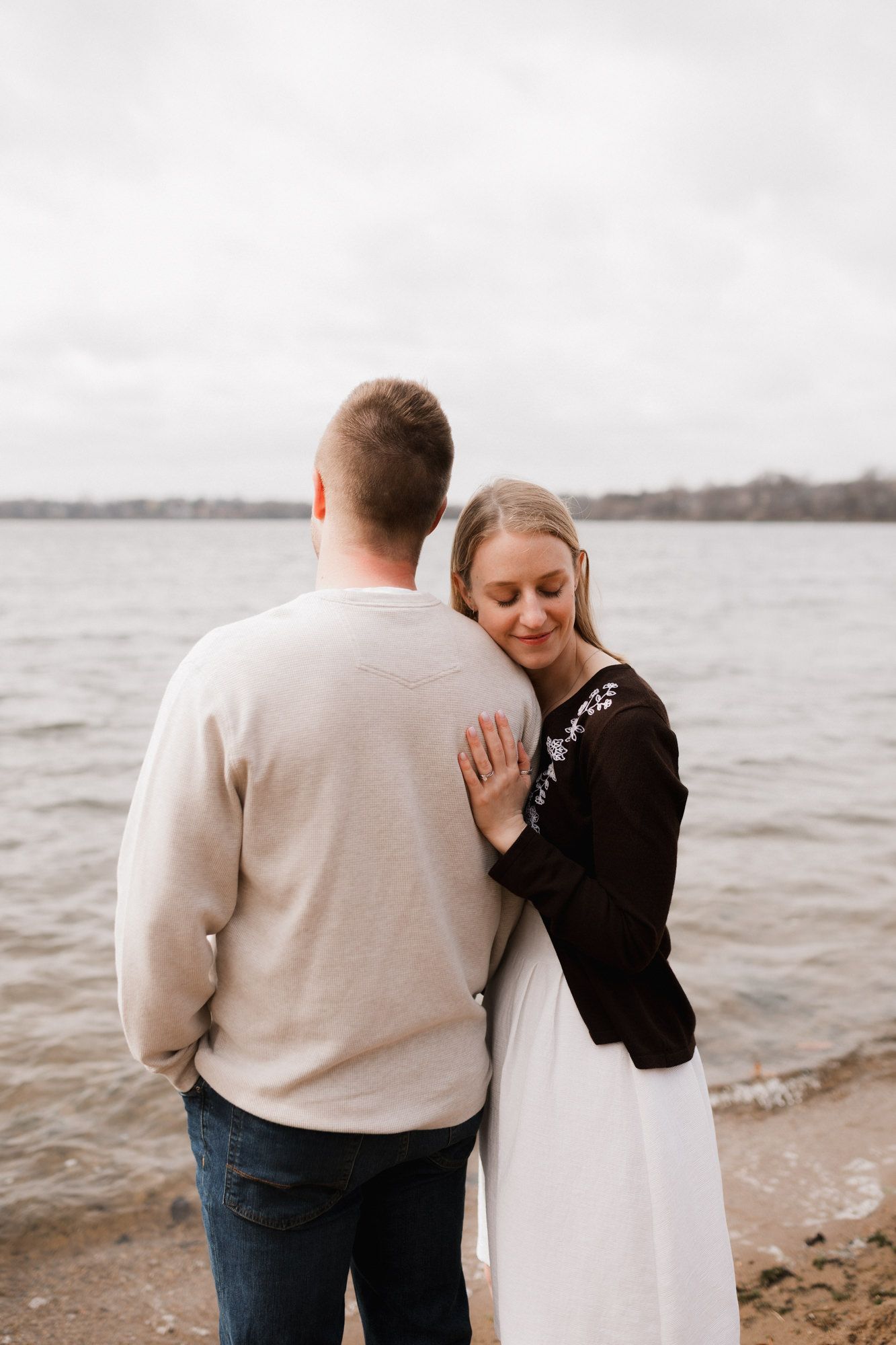 A woman rests her head on a mans shoulder while embracing near a lake on a cloudy day.