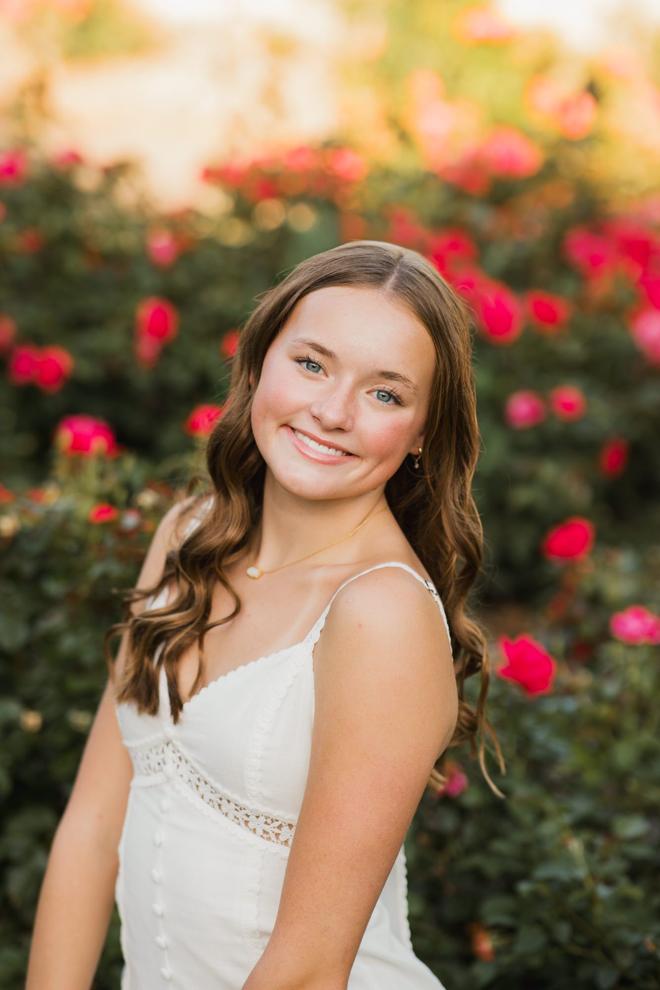 Young woman in white dress smiling in front of blooming red rose bushes in a garden portrait.
