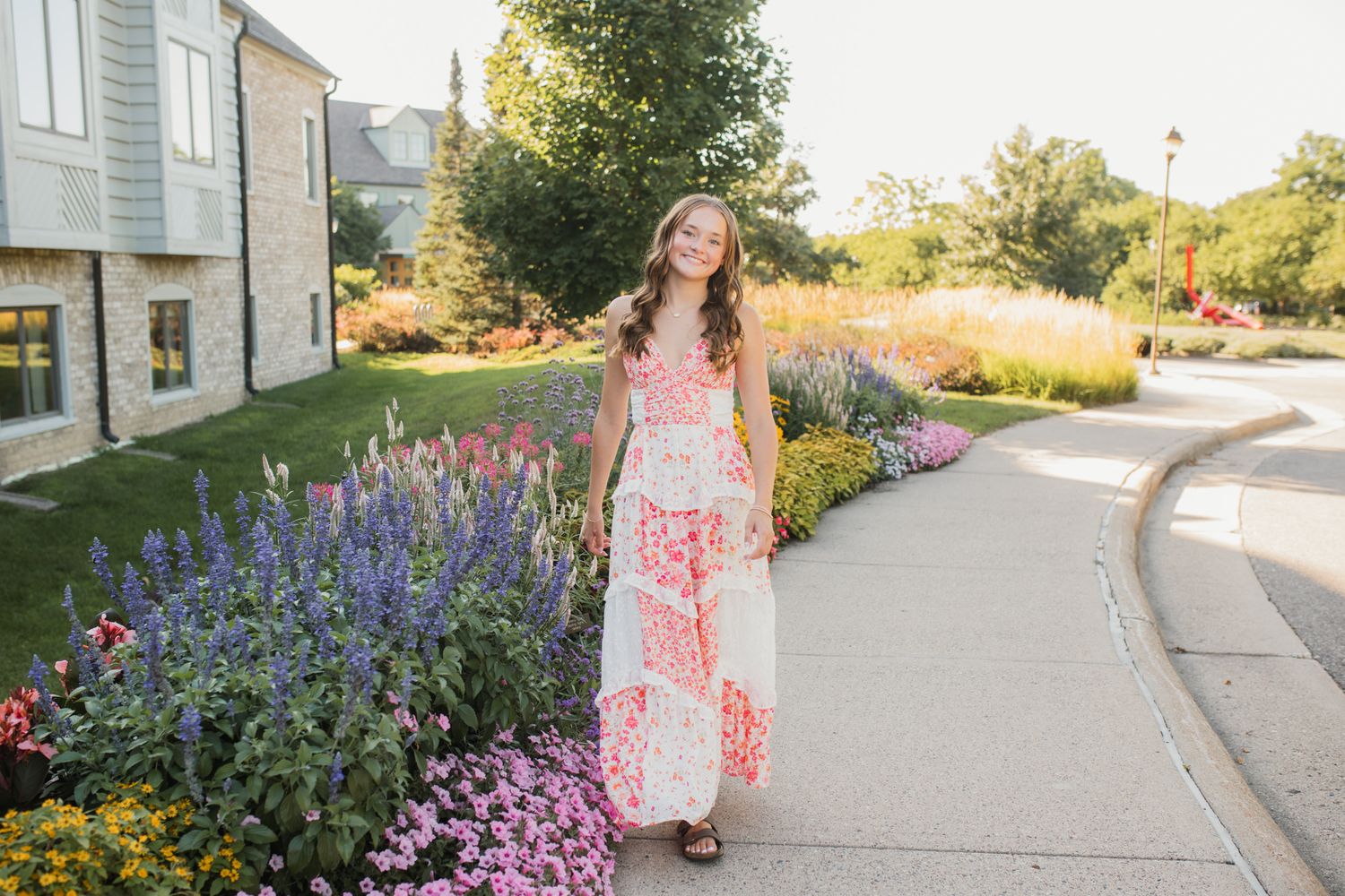 Young woman in floral maxi dress smiling on garden pathway surrounded by colorful blooming flowers on sunny day.