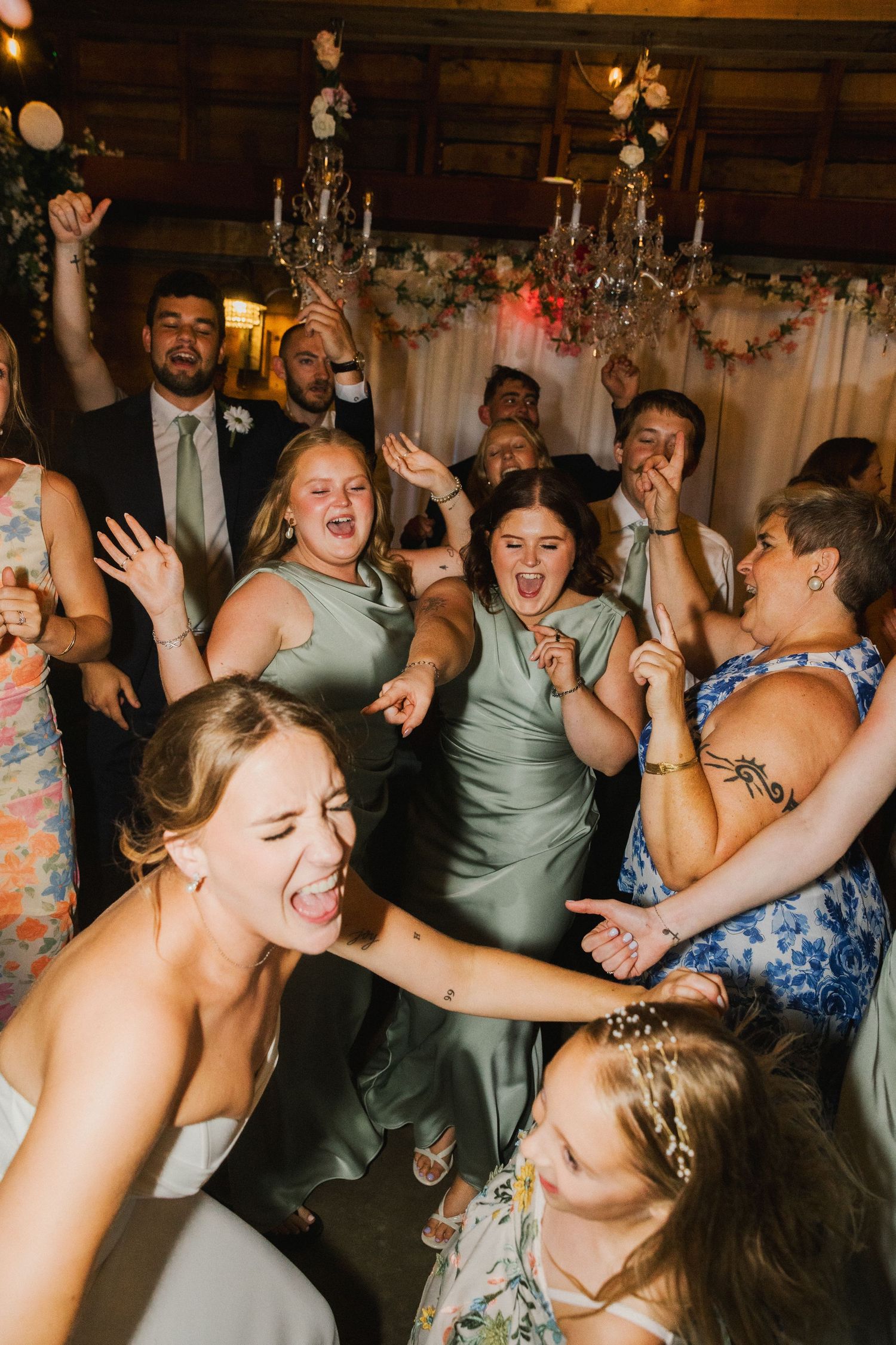 Excited wedding guests in sage green bridesmaid dresses dancing and celebrating joyfully at a rustic indoor wedding reception.